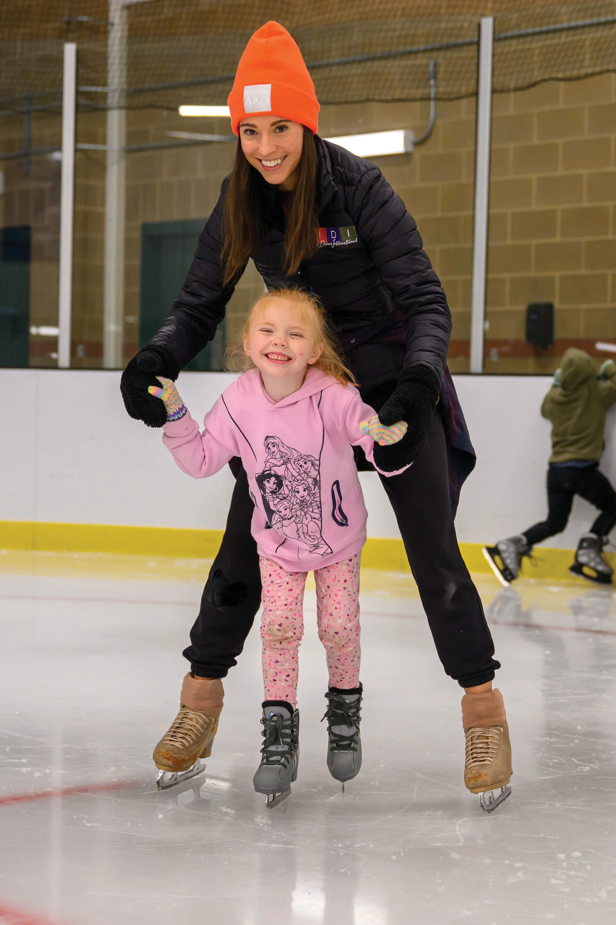 Ice skater in orange hat guiding a young skater in front of her.