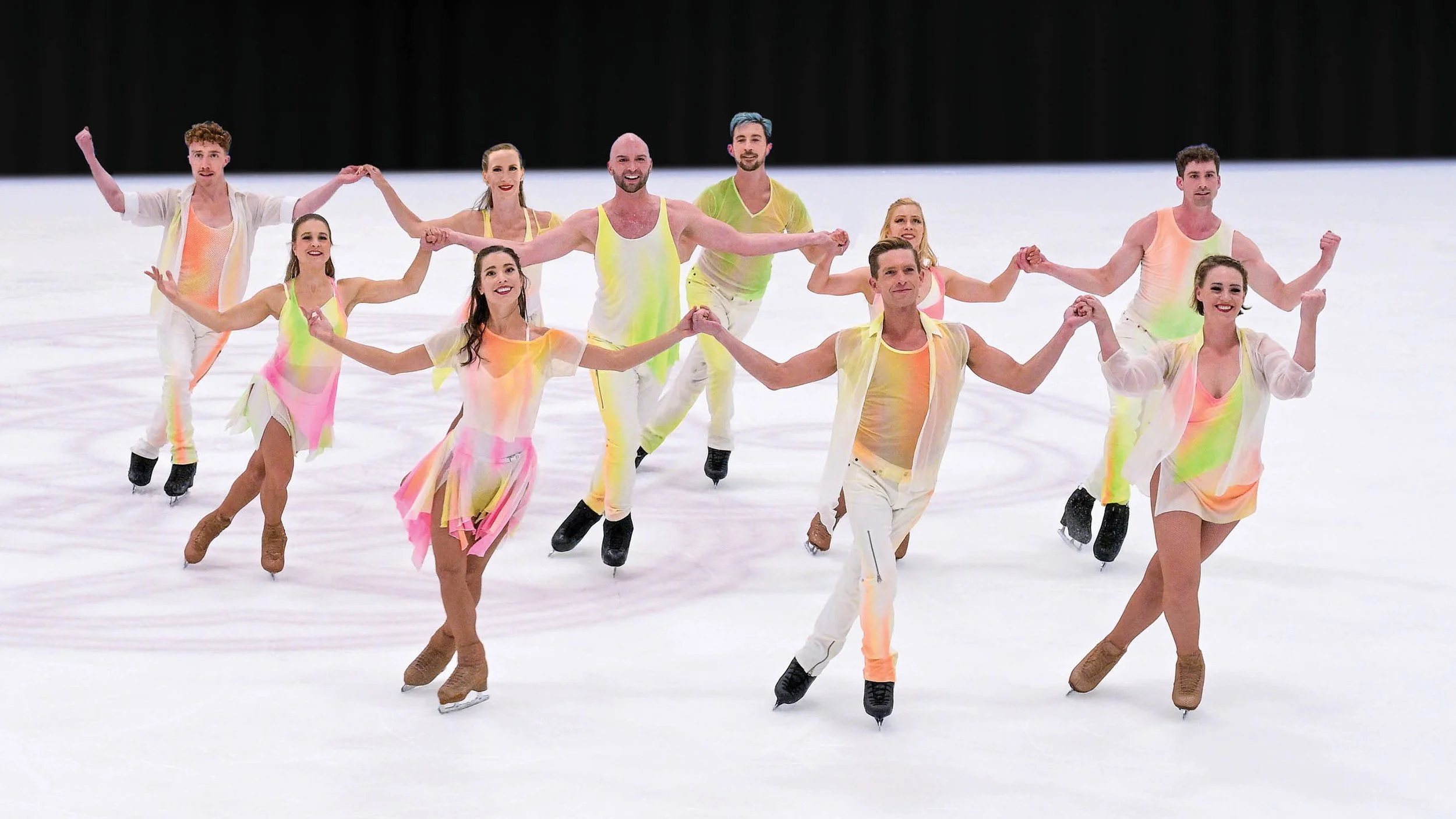 Ice skaters holding hands in rows in brightly colored costumes.