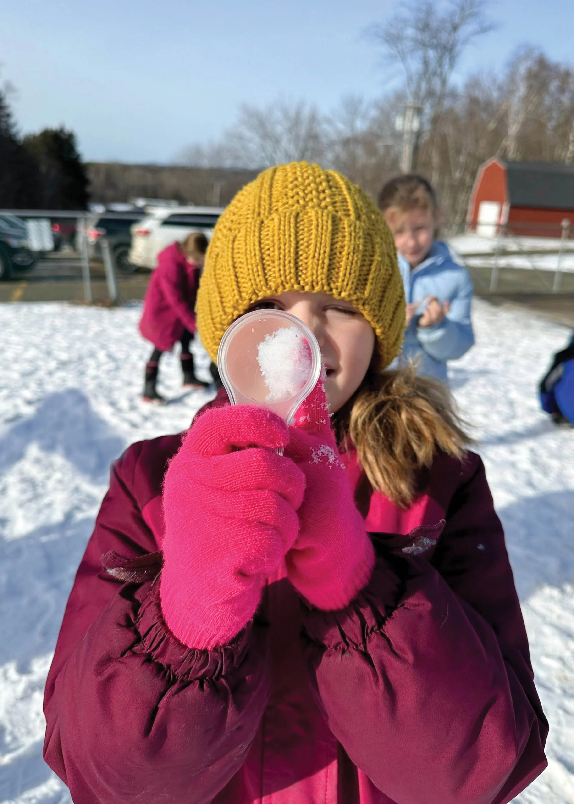 Girl with yellow hat holding a magnifying glass on snowy playground.