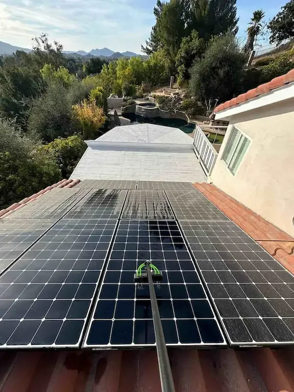 Close-up of solar panels on a roof with a hose cleaning the surface, overlooking a backyard with a swimming pool, trees, and mountains in the distance.