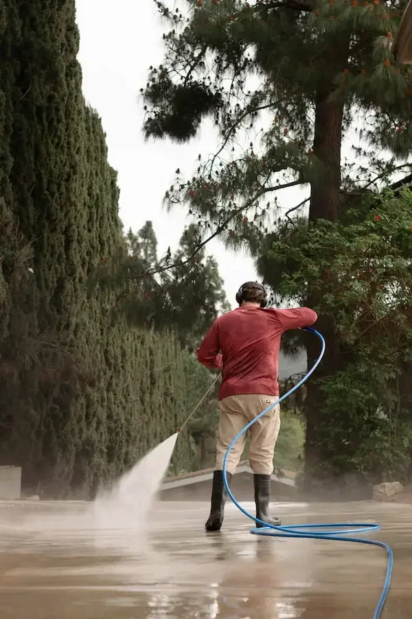 A man wearing a red shirt, beige pants, and black boots uses a hose to spray water on a large concrete surface surrounded by tall trees.