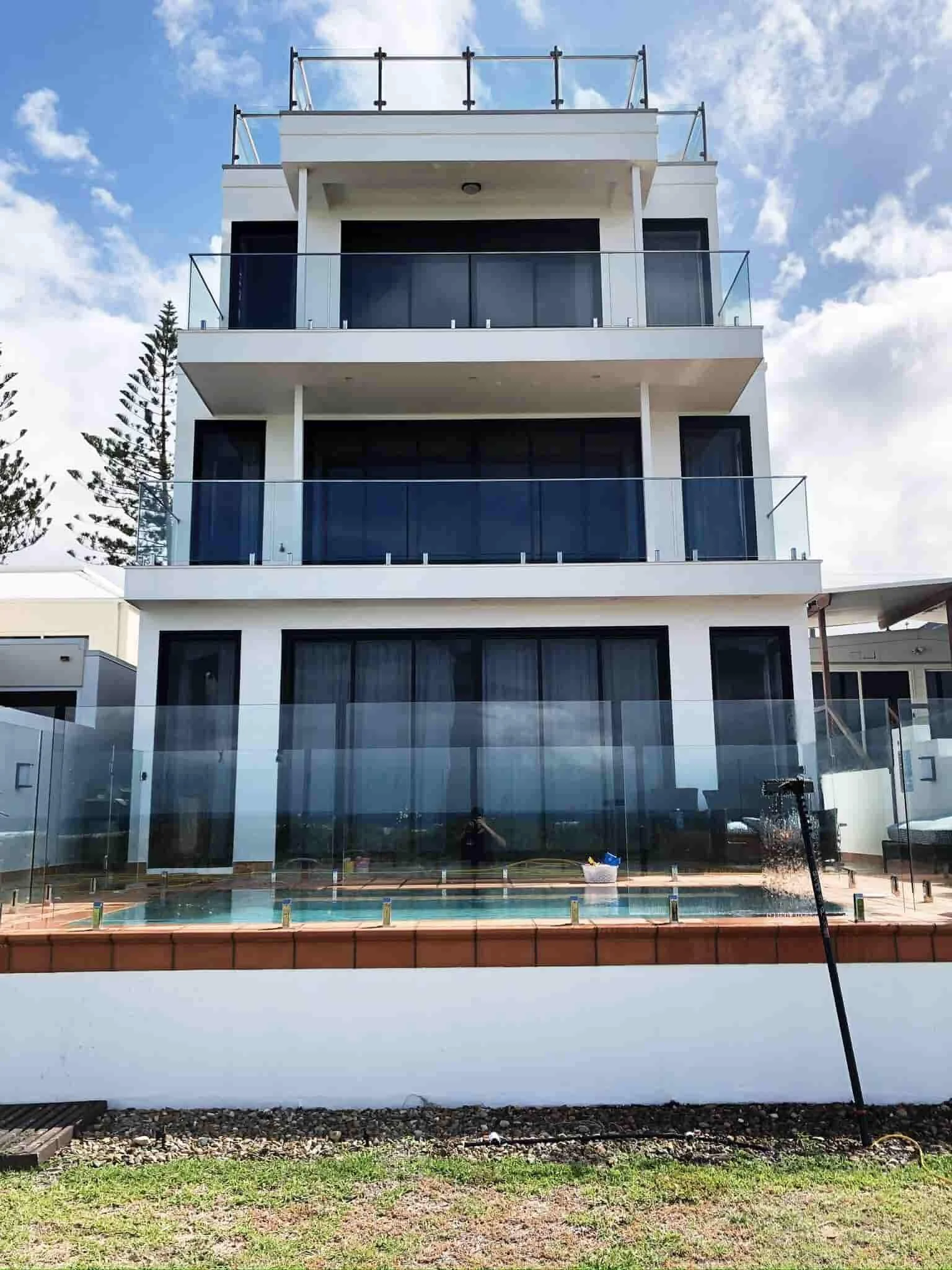 Modern white building with three floors, each with glass balconies, large windows, and a rooftop deck, against a partly cloudy sky.