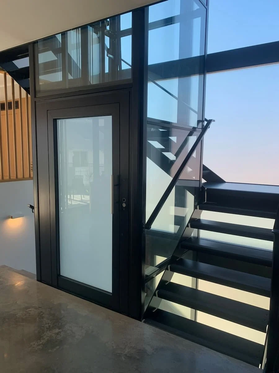 Modern interior with a black staircase and glass elevator shaft near a large window with a city view, polished concrete floor, and wooden wall panels.