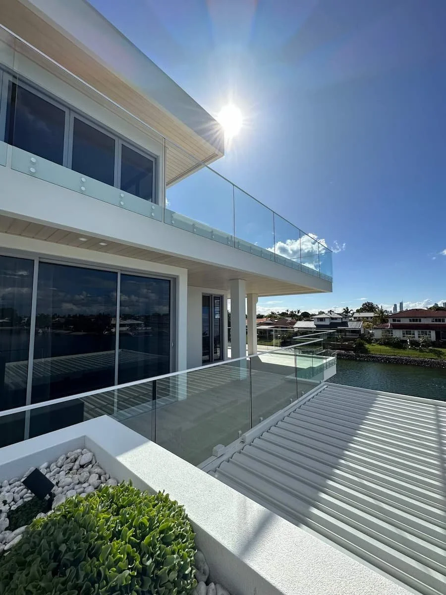 Modern waterfront building with glass balcony railings, large windows, and a rooftop patio under a bright sun in the clear blue sky.