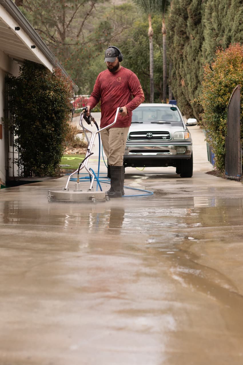 A man with a beard wearing a cap, red long-sleeve shirt, and beige pants operating a pressure washer to clean a concrete driveway, with a parked car nearby.