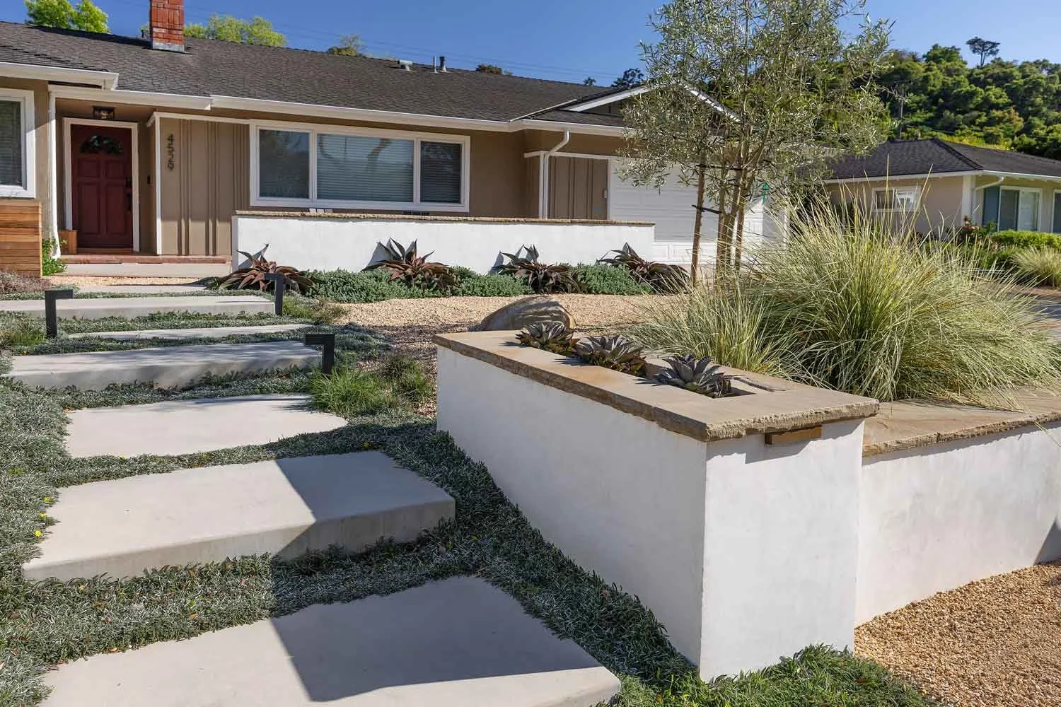 Modern front yard with succulent and grass landscaping, paved walkway, and single-story house with beige exterior and red front door.