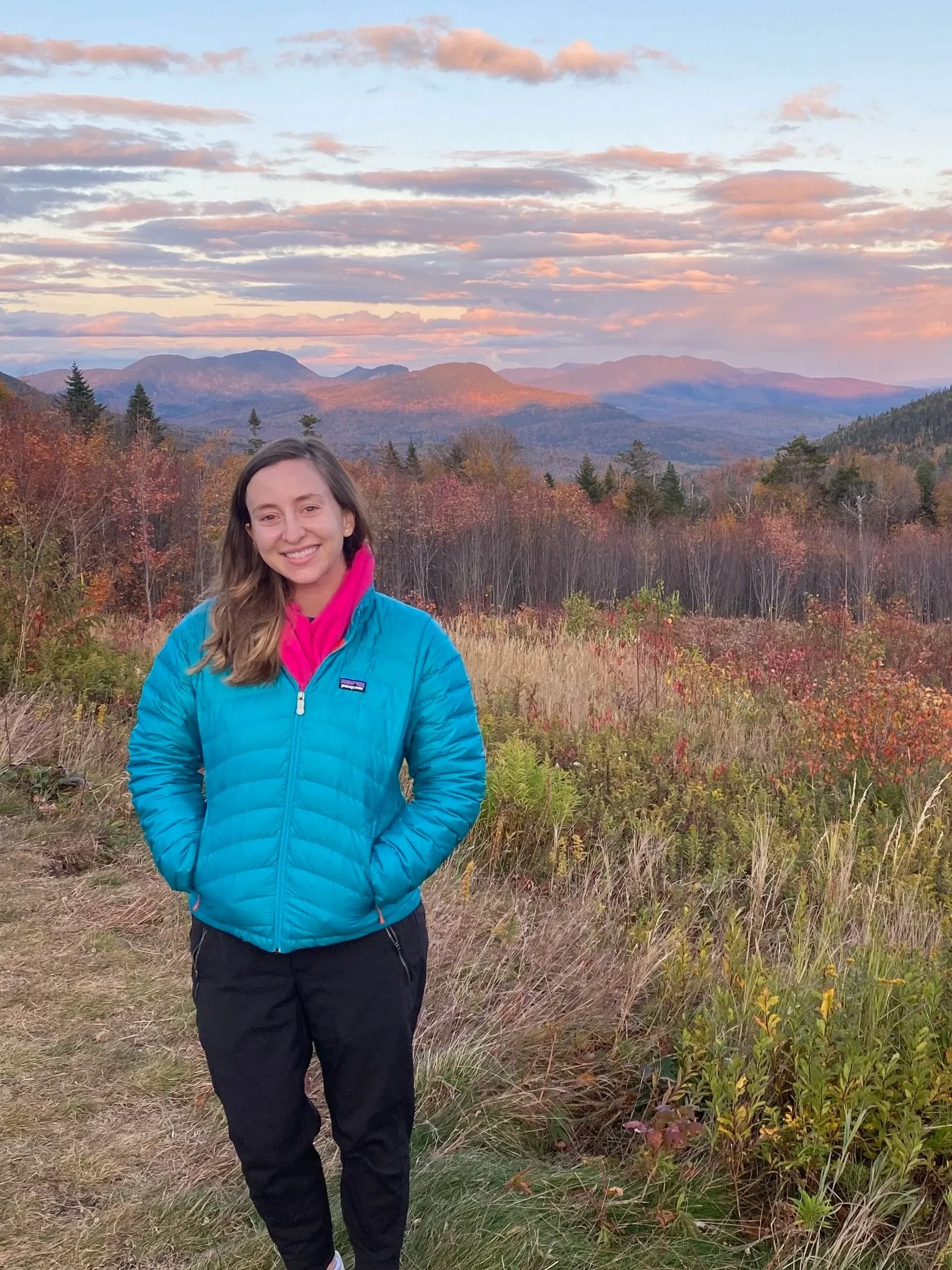 A smiling woman wearing a blue jacket and black pants standing in a grassy area with colorful fall foliage, trees, mountains, and a partly cloudy sky in the background during sunset.