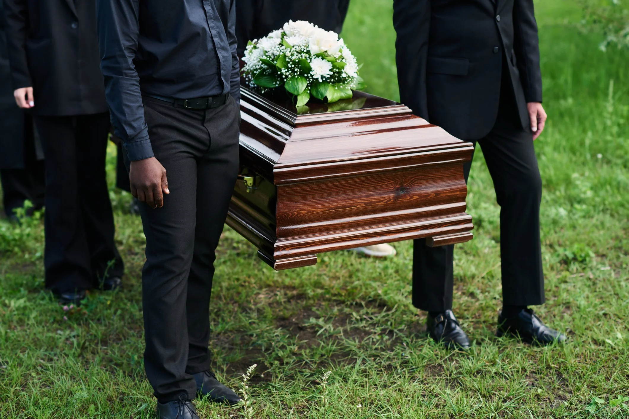 Pallbearers carrying a wooden casket with flowers on top in a grassy area, dressed in formal black attire.