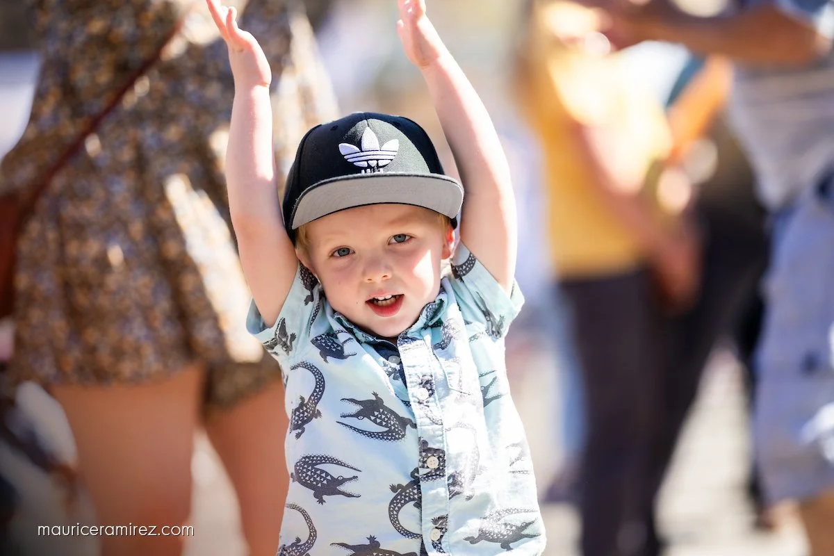 Young boy with light hair, wearing a baseball cap and a shirt with dinosaur patterns, raising his arms outdoors with adults in the background.