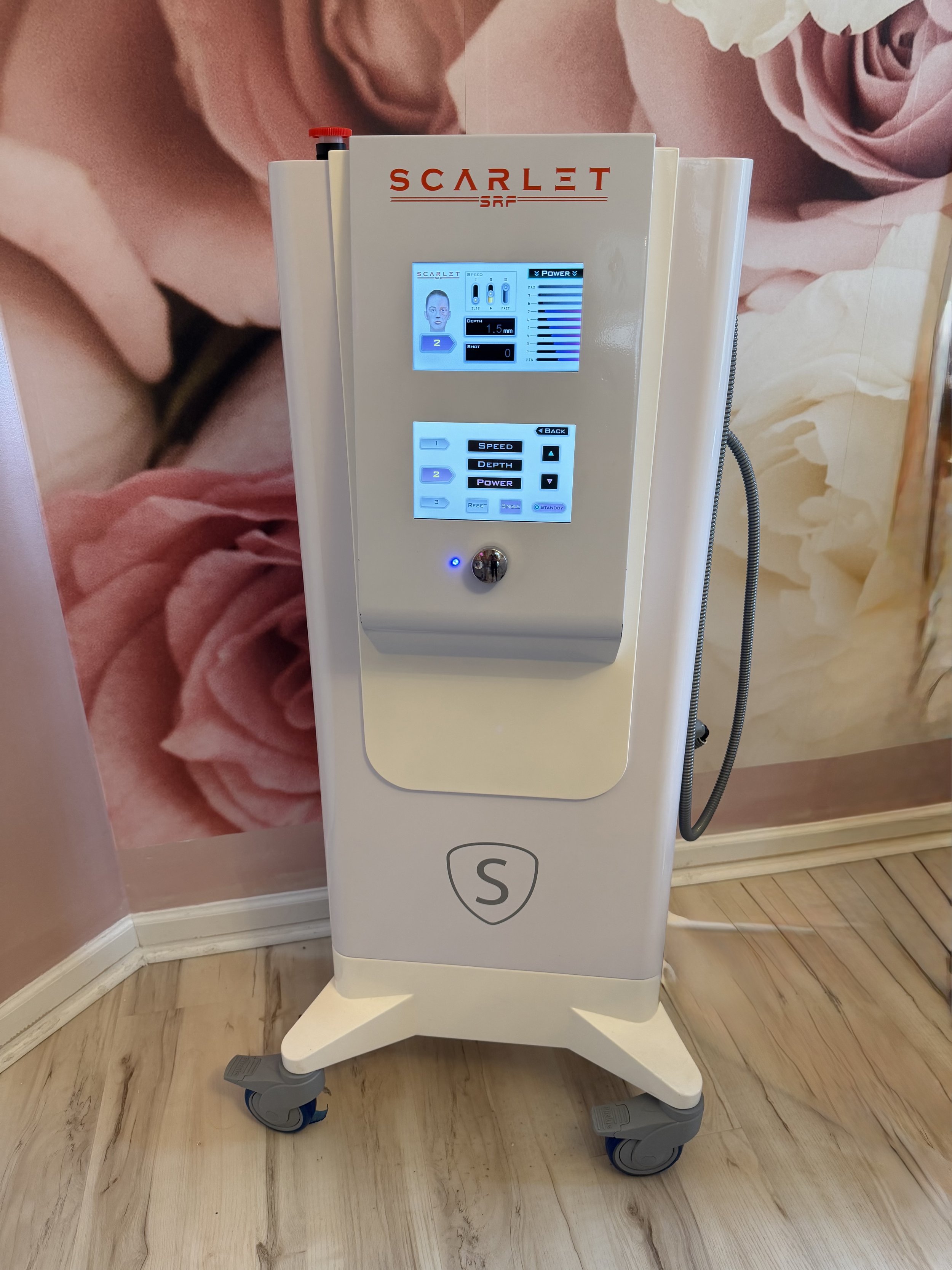 Medical or aesthetic device on a mobile stand with a digital interface, display screen, and control buttons. Background features a large floral mural wall. Wooden floor.