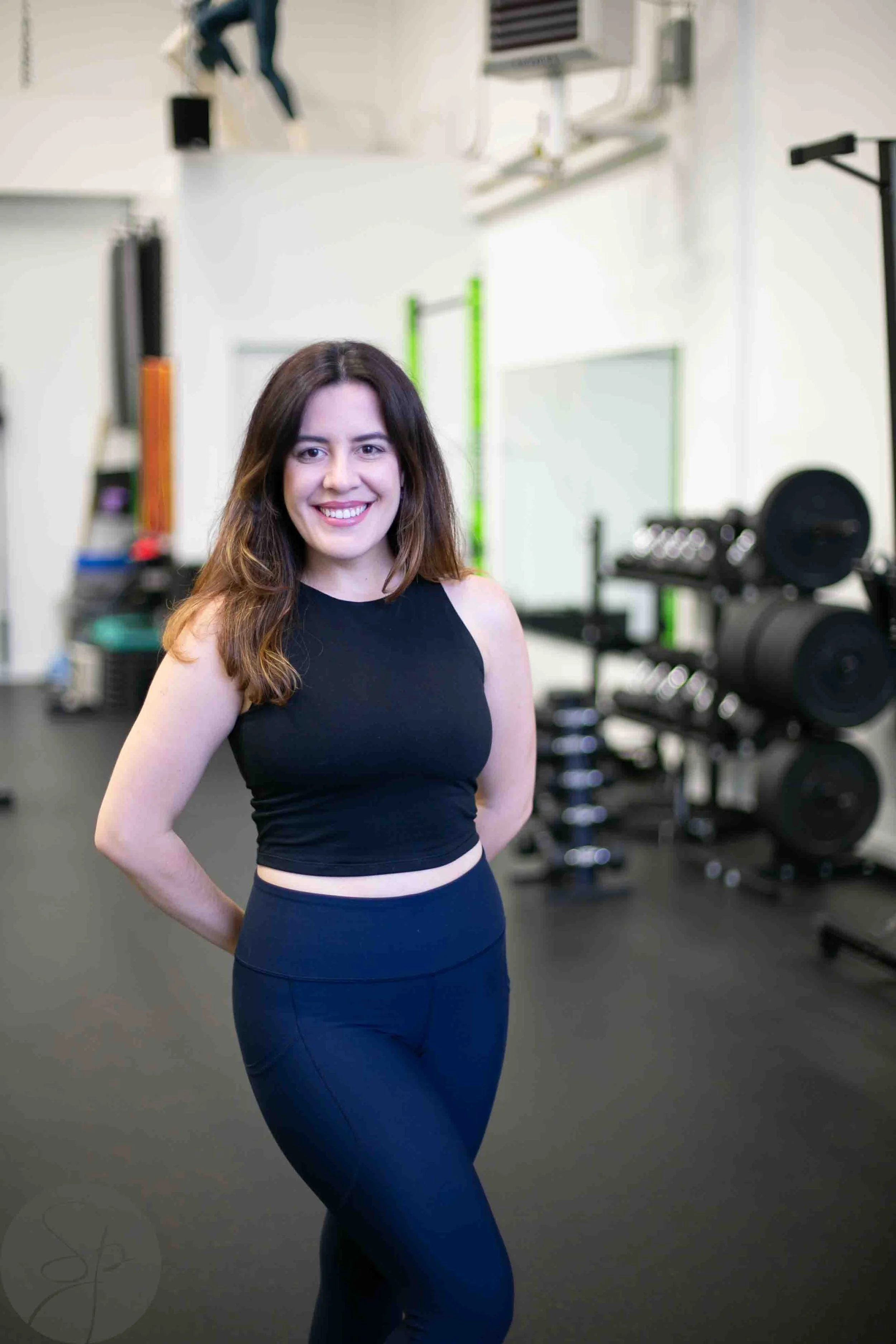 A woman in a black workout outfit standing in a gym with exercise equipment in the background.