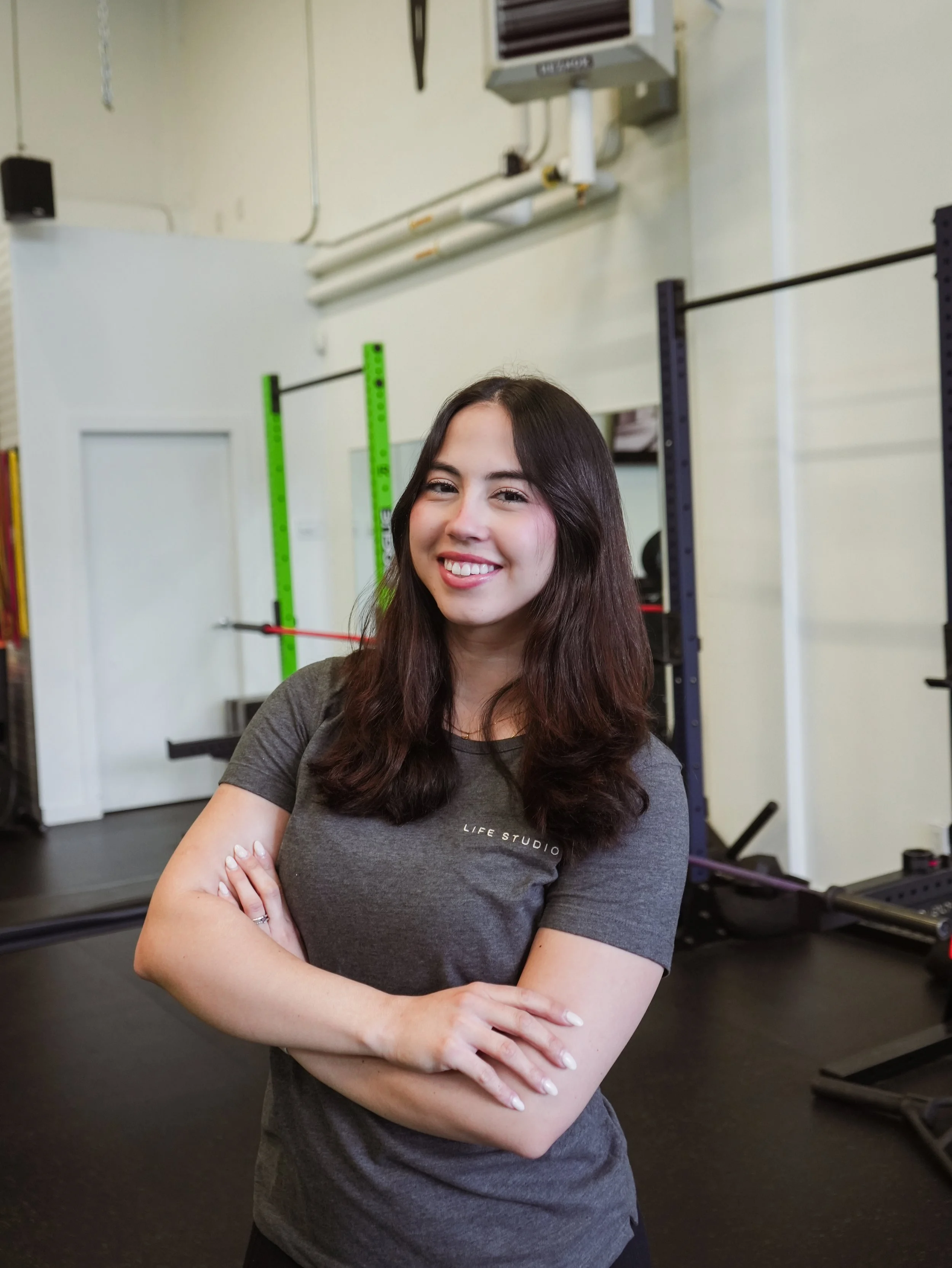Gabriella smiling, standing with arms crossed in a gym setting with exercise equipment in the background.