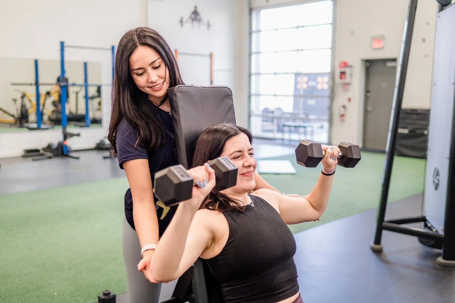 Two women in a gym, one lifting dumbbells while seated, with the other assisting. They are surrounded by gym equipment and bright natural light.