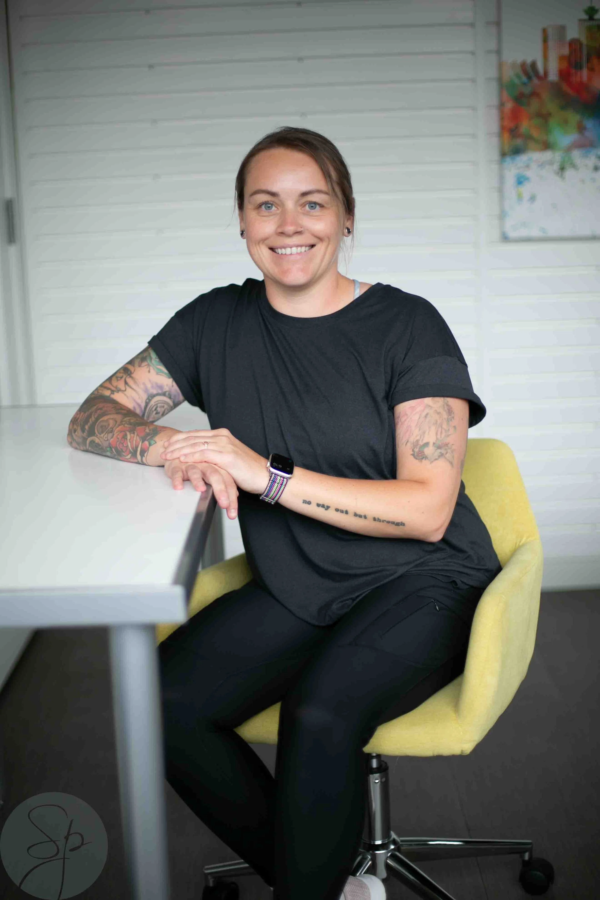 Person with tattoos sitting on a yellow chair, smiling, wearing a black shirt and a smartwatch.