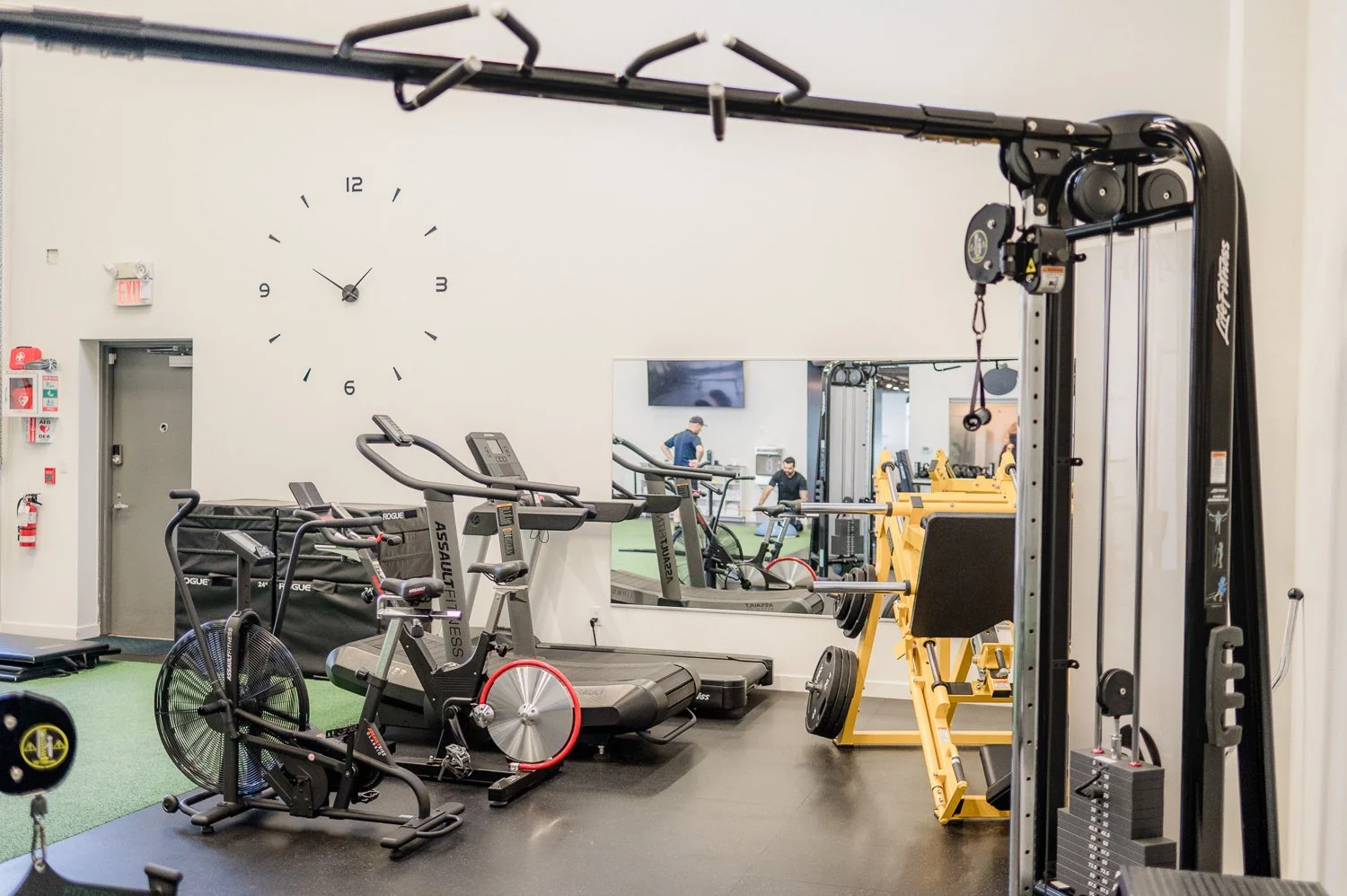 Modern gym interior with exercise equipment, including a treadmill, stationary bike, weight machine, and wall clock.