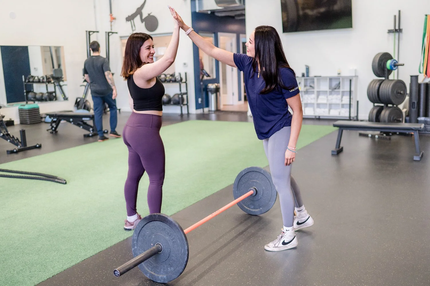 Two women high-fiving in a gym beside a barbell, with workout equipment in the background.