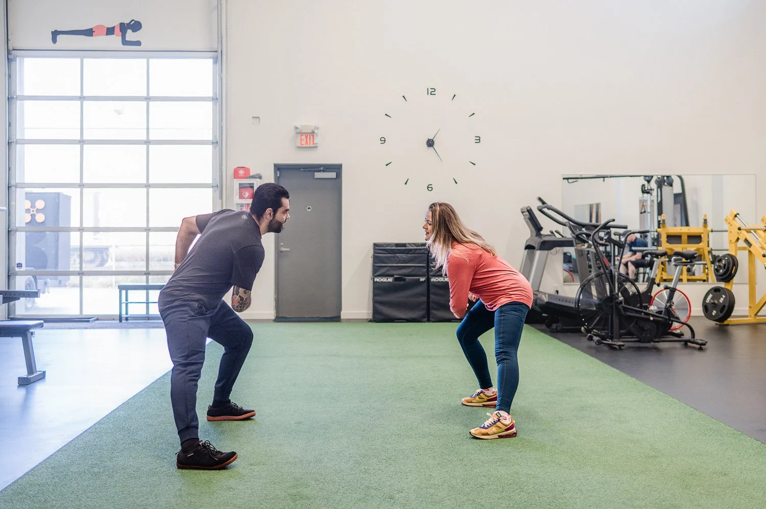 Two people practicing squats in a gym with exercise equipment and a wall clock.