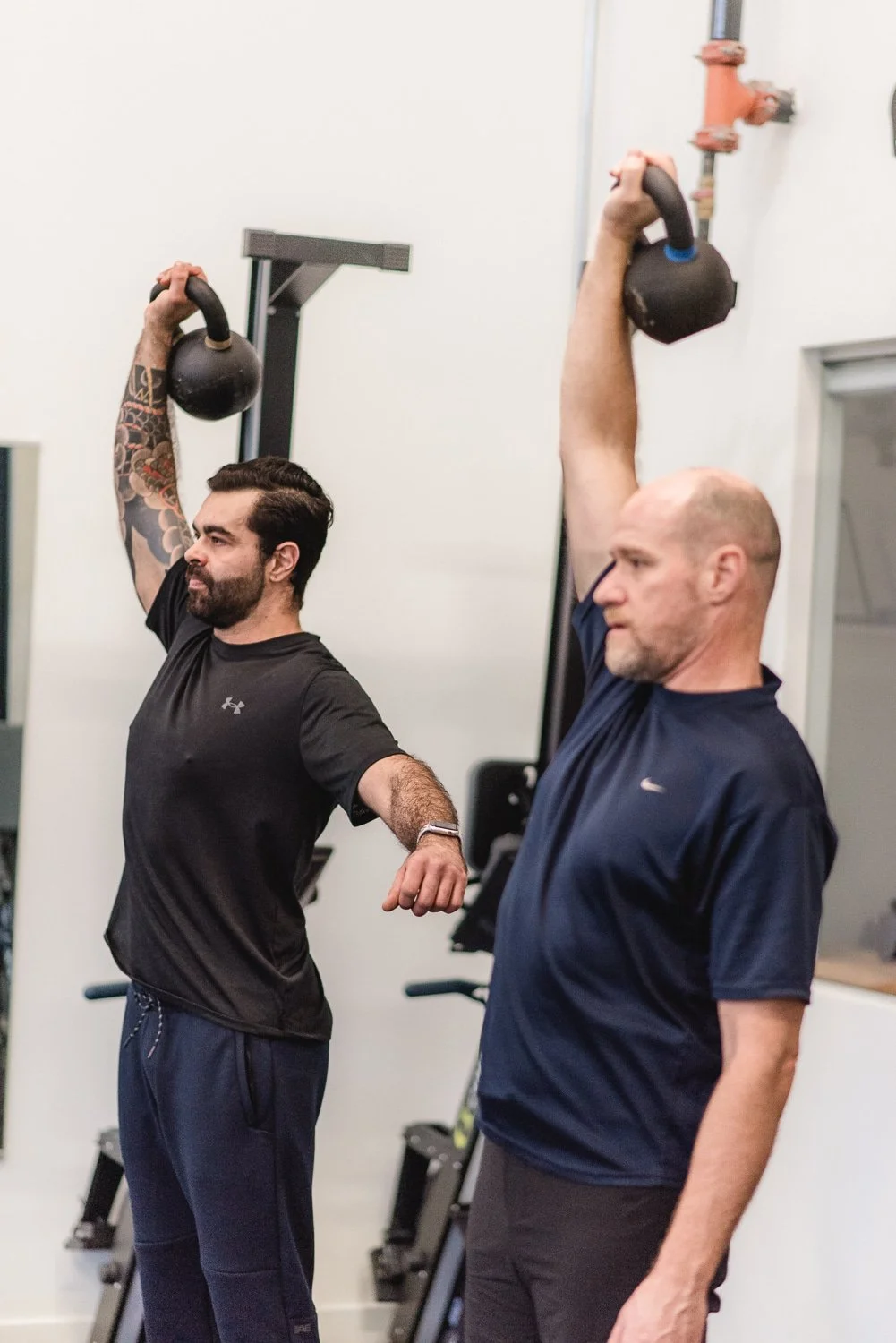 Two men in athletic clothing doing kettlebell exercises indoors, raising one arm each overhead, concentrating on form.