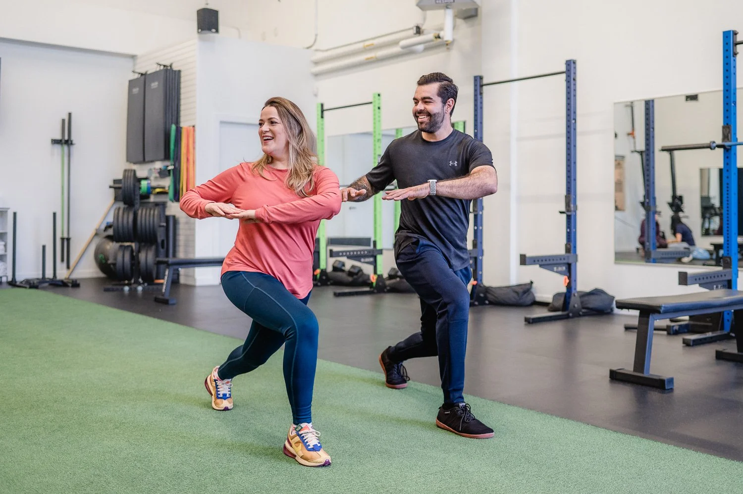 Two people exercising in a gym, performing lunges with their arms outstretched, surrounded by fitness equipment.