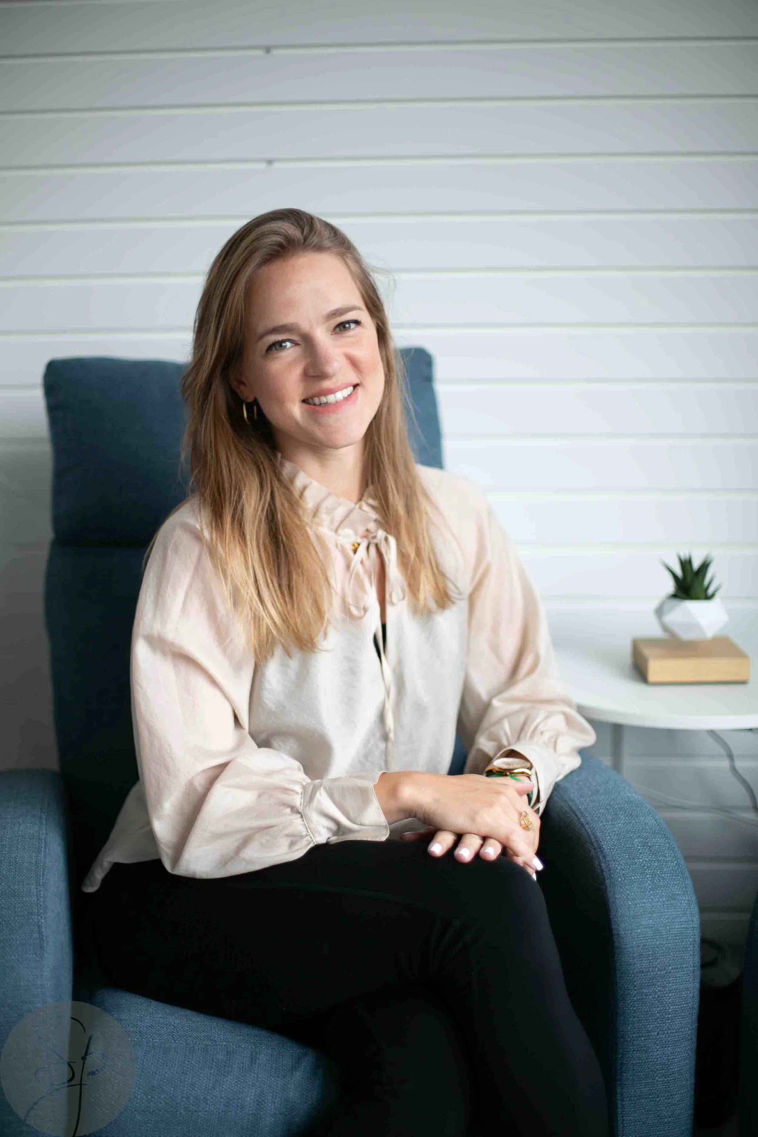 Woman sitting in a blue chair, smiling, wearing a light blouse, with white wall background.