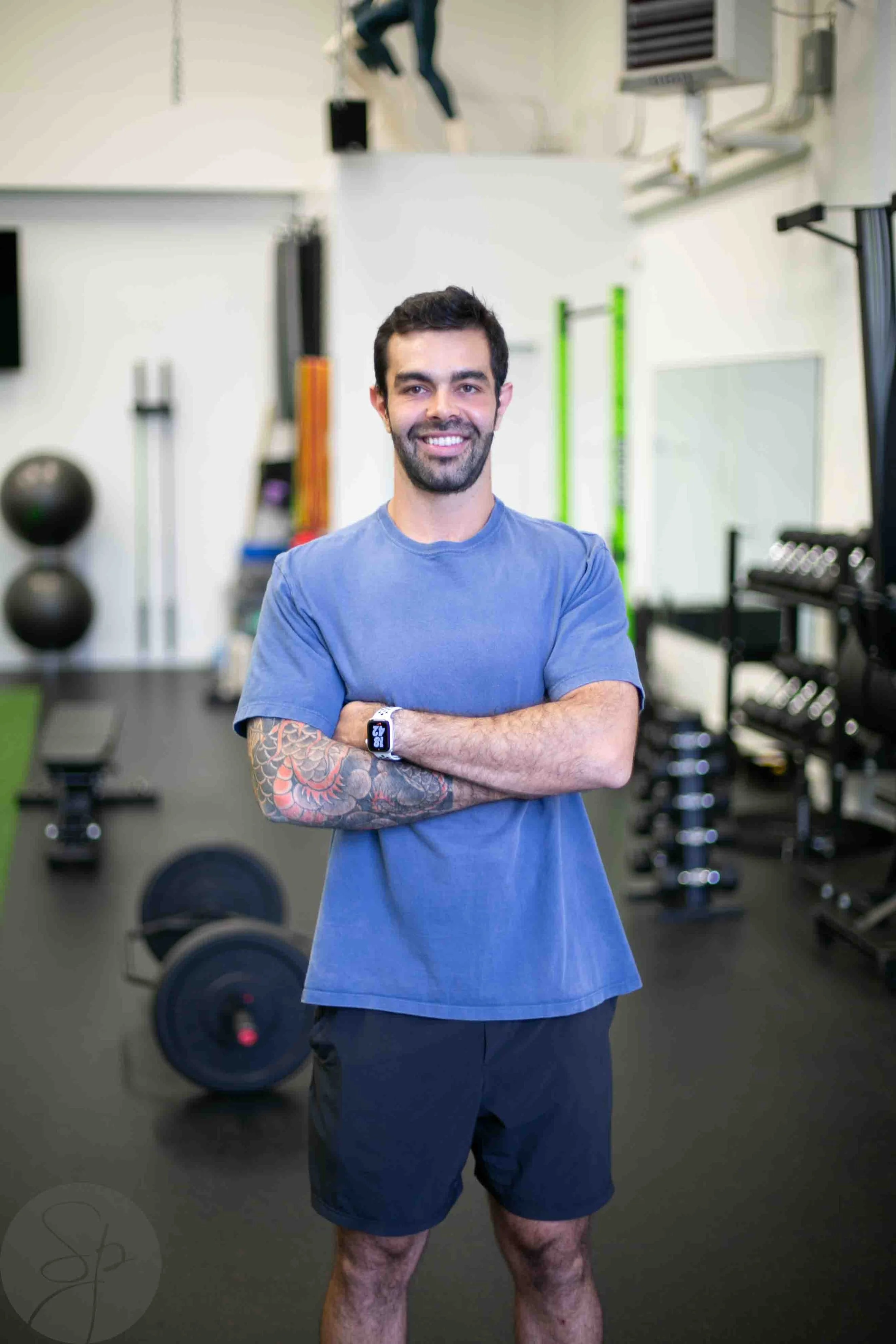 Man with tattoos and a smartwatch standing in a gym, exercising equipment in the background.