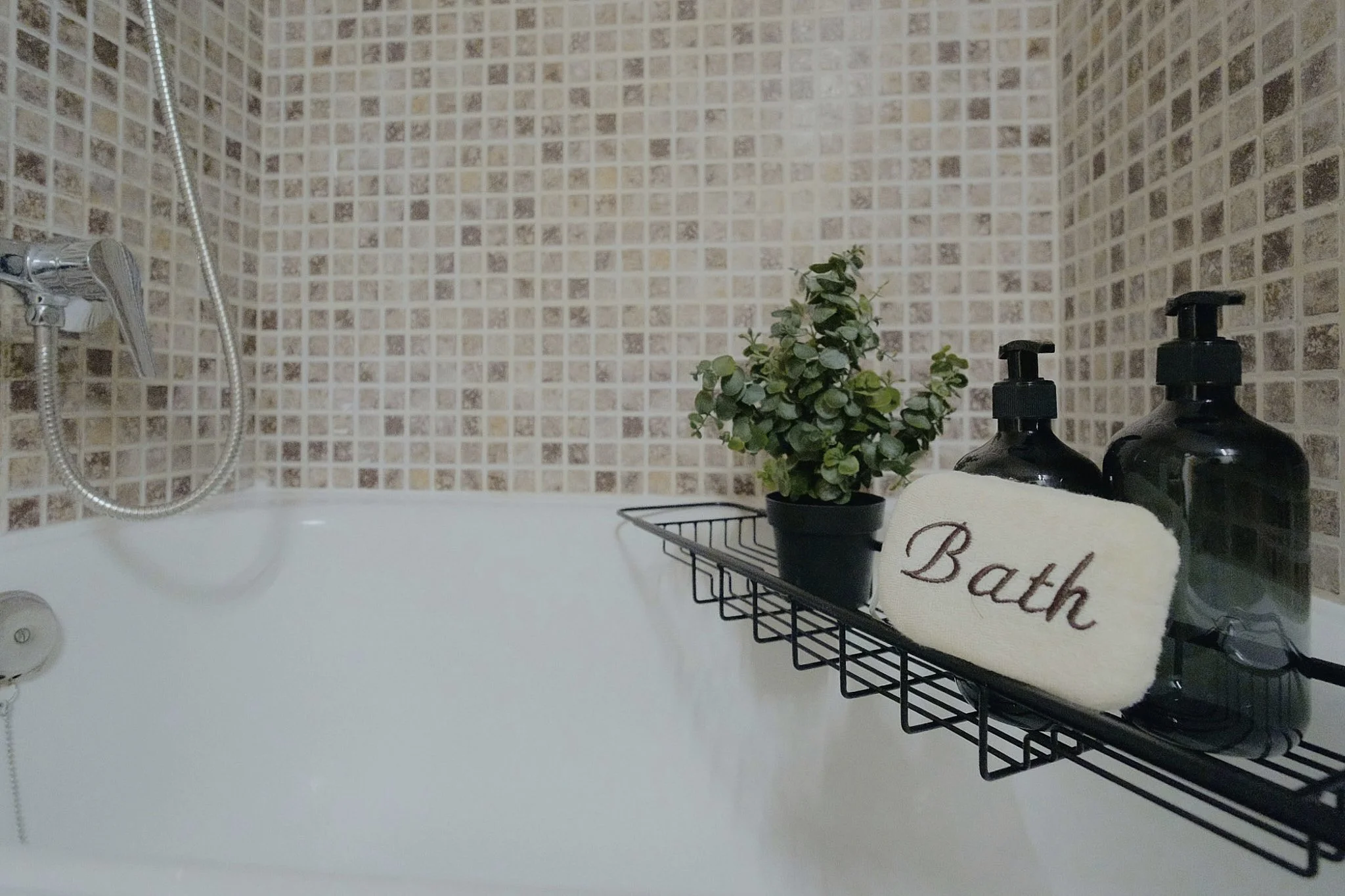 Una bañera con una pared de azulejos en tonos beige y marrón. Sobre la bañera, hay una cesta de metal con dos botellas negras y un toallero que dice 'Bath', con una planta y un bote de jabón o loción.