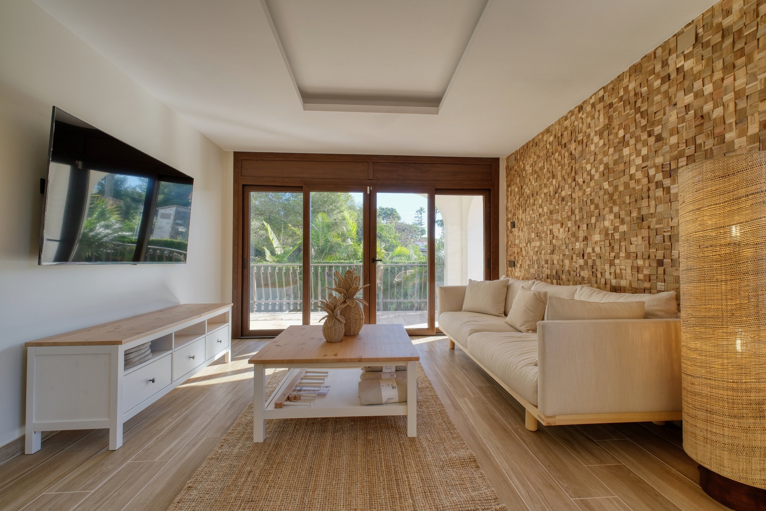 Sala de estar con pared de madera y ventana grande con vista a árboles, sofá beige, mesa de centro blanca y lámpara de mimbre.