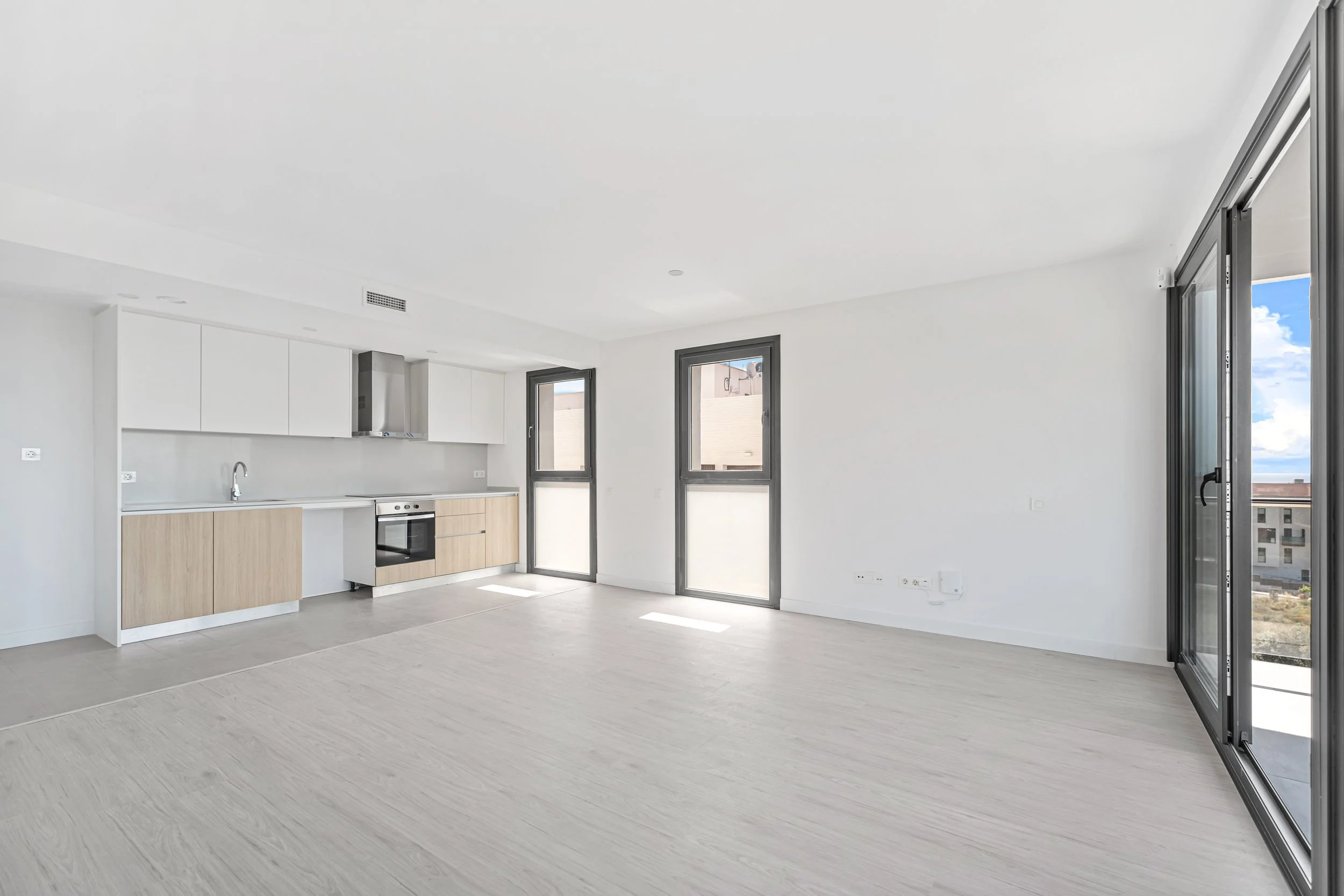 Interior de un departamento vacío con cocina moderna, ventanas grandes y balcón con vista al cielo y edificios