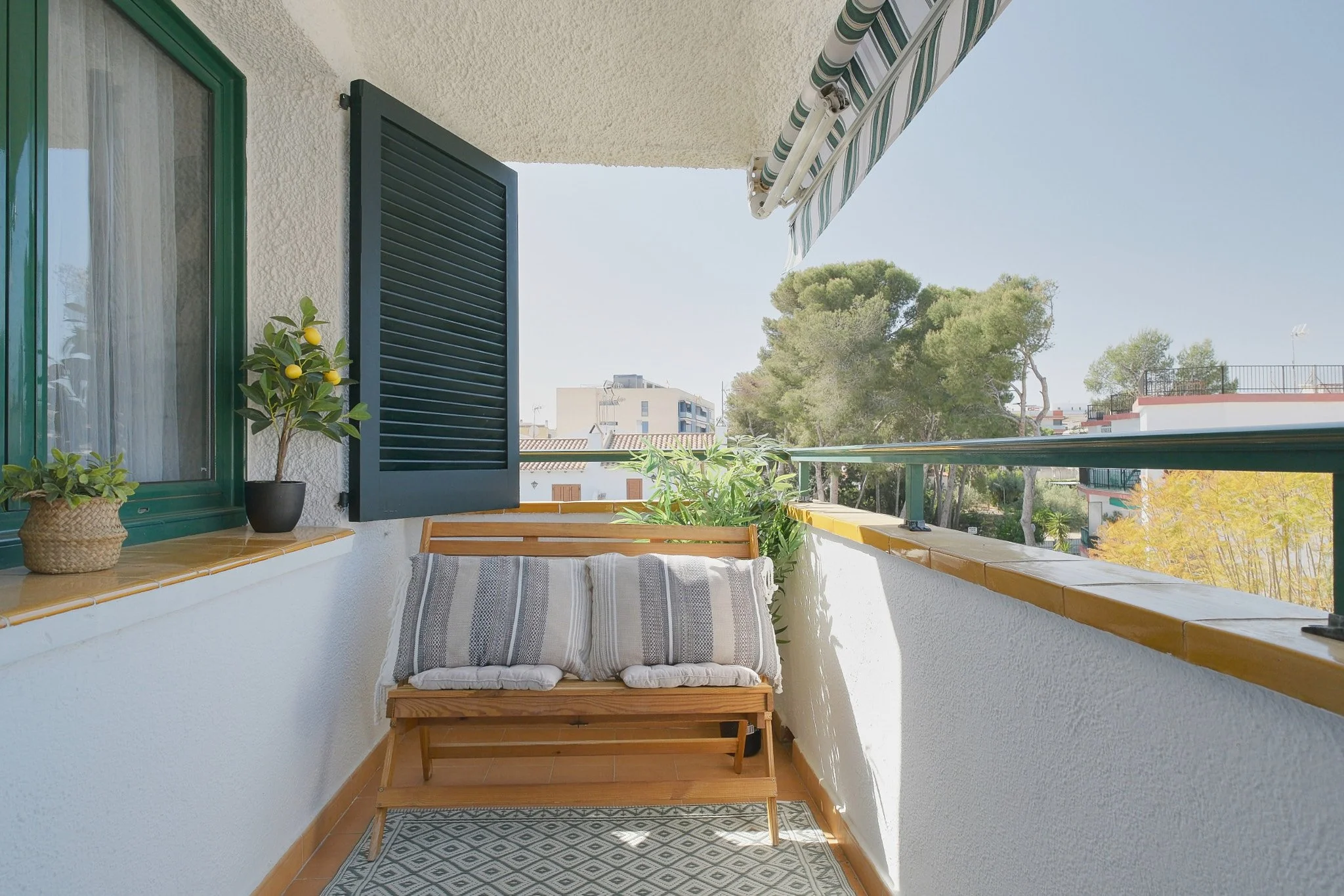 Vista de un balcón con mueble de madera, cojines, plantas en macetas y vista hacia árboles y edificios en un día soleado.