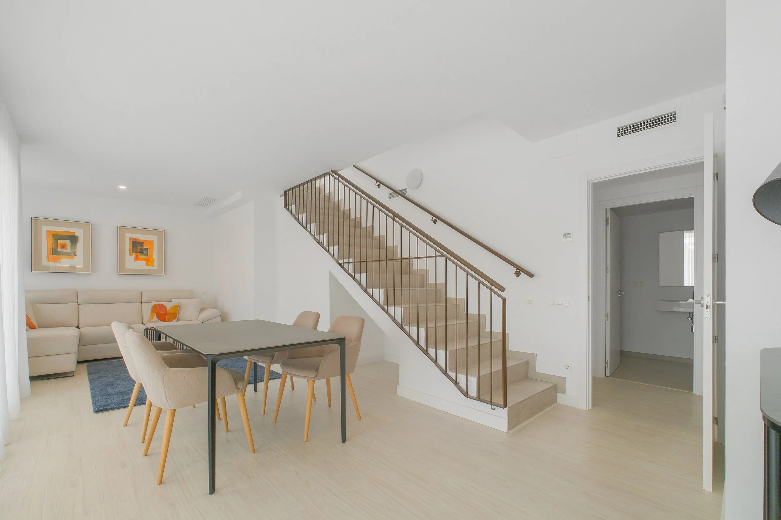 Sala moderna con sofá beige, mesas y sillas de comedor de madera, paredes blancas con arte abstracto, escalera de madera con barandal de metal, y puerta blanca en fondo.