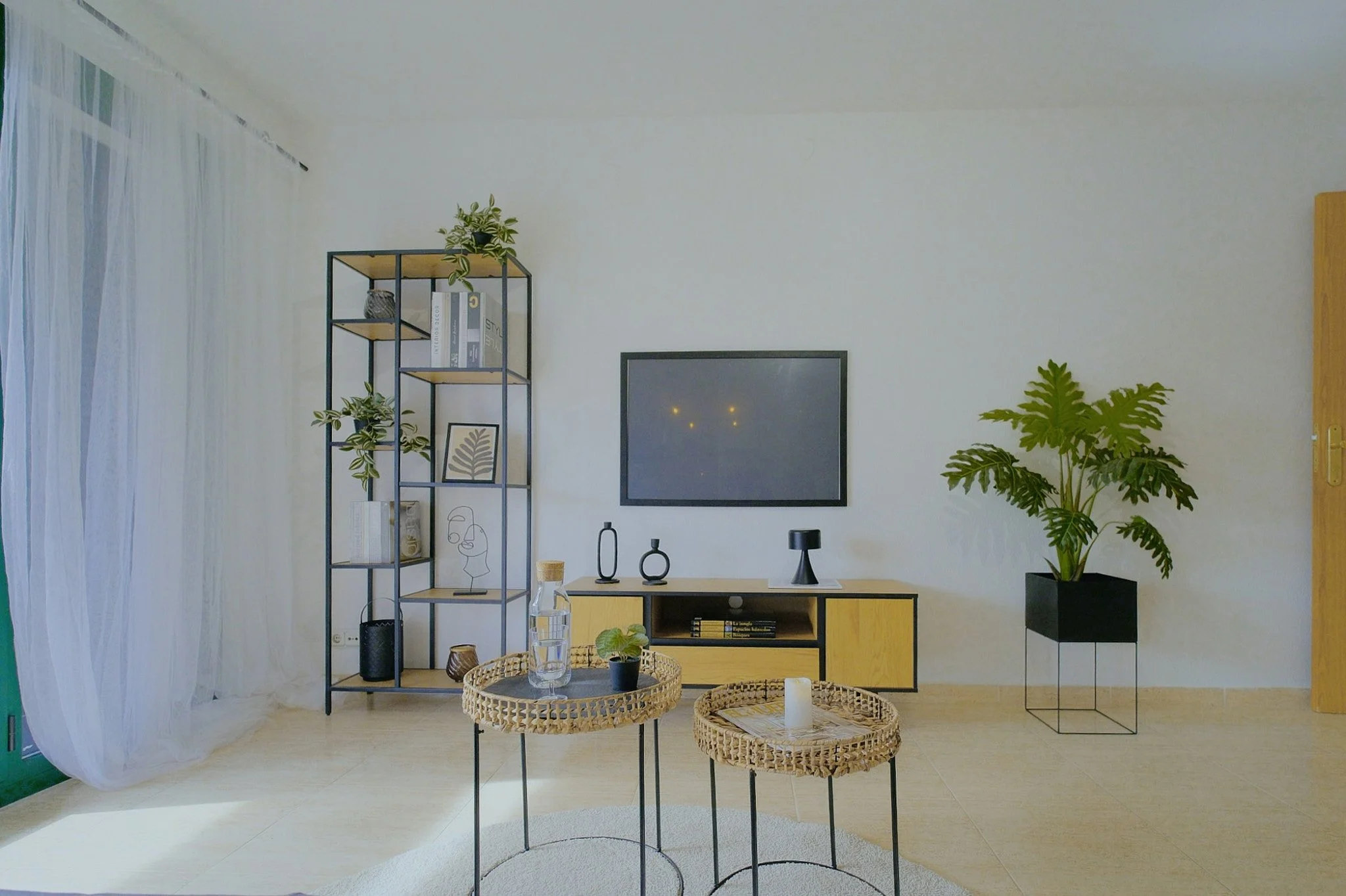 Interior de una sala con estantería negra, plantas y decoraciones minimalistas. Tiene una pared blanca, cortinas blancas al lado izquierdo, y una mesa con jarrones y libros.