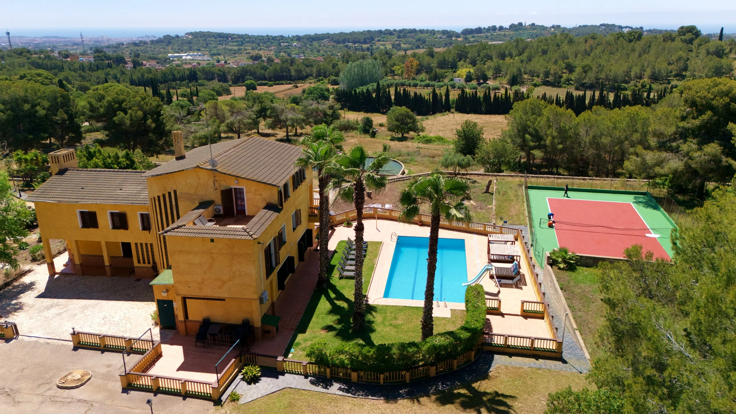 Vista aérea de una casa de estilo mediterráneo con piscina, palmeras y cancha de tenis, de lujo, de alquiler turistico en vilanova y la geltrú