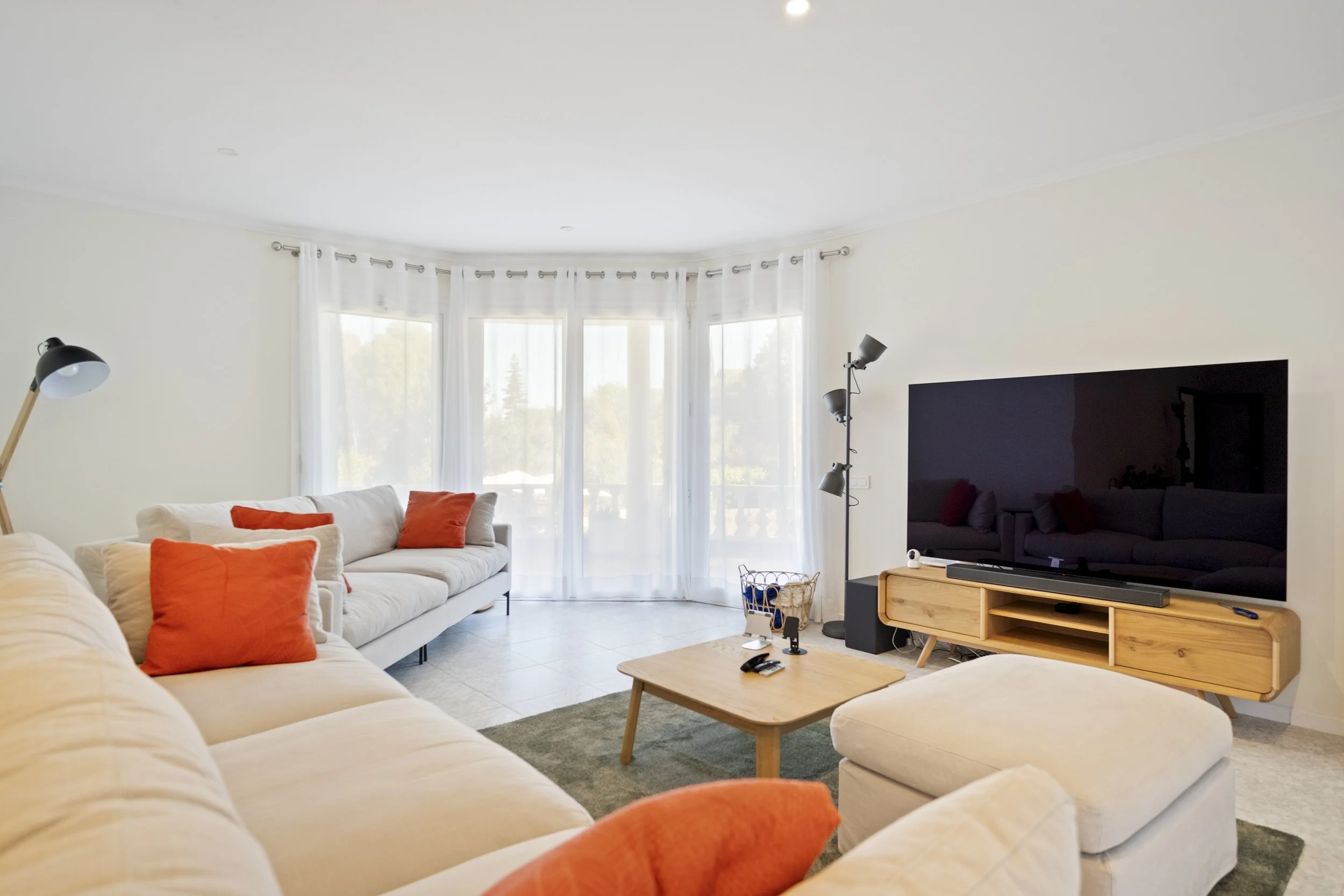 Sala de estar con sillones blancos con cojines naranjas, una televisión grande en un mueble de madera, cortinas blancas y una ventana grande que permite mucha luz natural, una lámpara de pie con varias y una mesa de centro de madera.