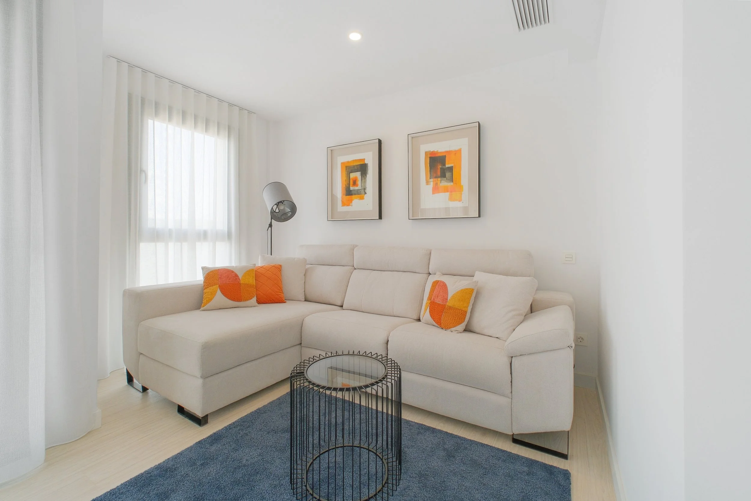 Sala moderna con sofá blanco y cojines naranjas y amarillos, ventanas con cortinas blancas, cuadros abstractos en la pared, lámpara de pie y mesa de centro de metal negro.