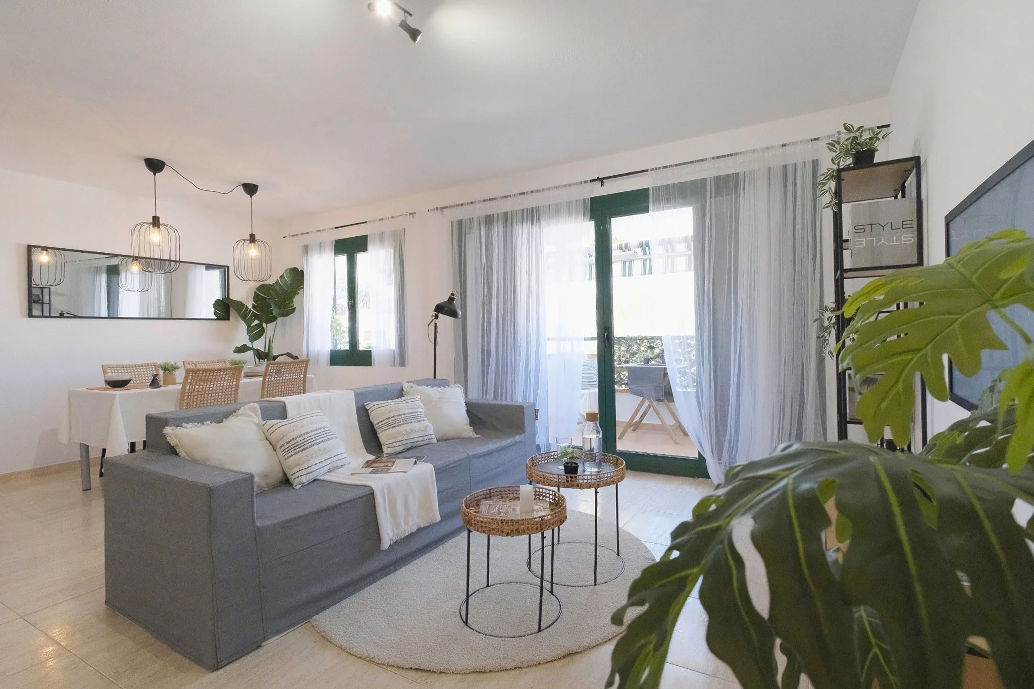 Sala de estar con sofá gris, mesa de centro de mimbre, cortinas blancas y ventana grande con vista a un balcón, plantas decorativas y lámparas de techo de estilo industrial.