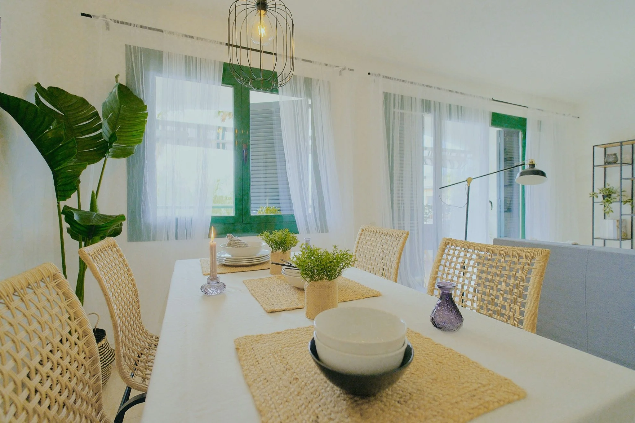 Interior de comedor con mesa decorada con plantas, velas y vajilla, rodeada de sillas de mimbre, ventanas grandes con cortinas blancas, lámpara de araña y una lámpara de pared.
