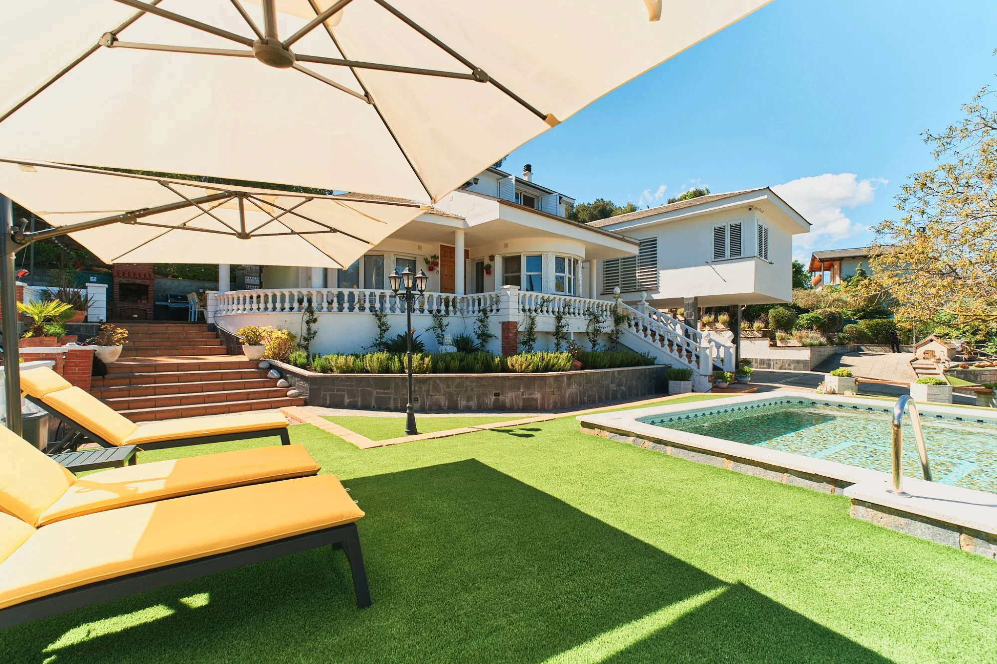 Casa blanca moderna con terraza, piscina y área de jardín con sombrillas y tumbonas en un día soleado.