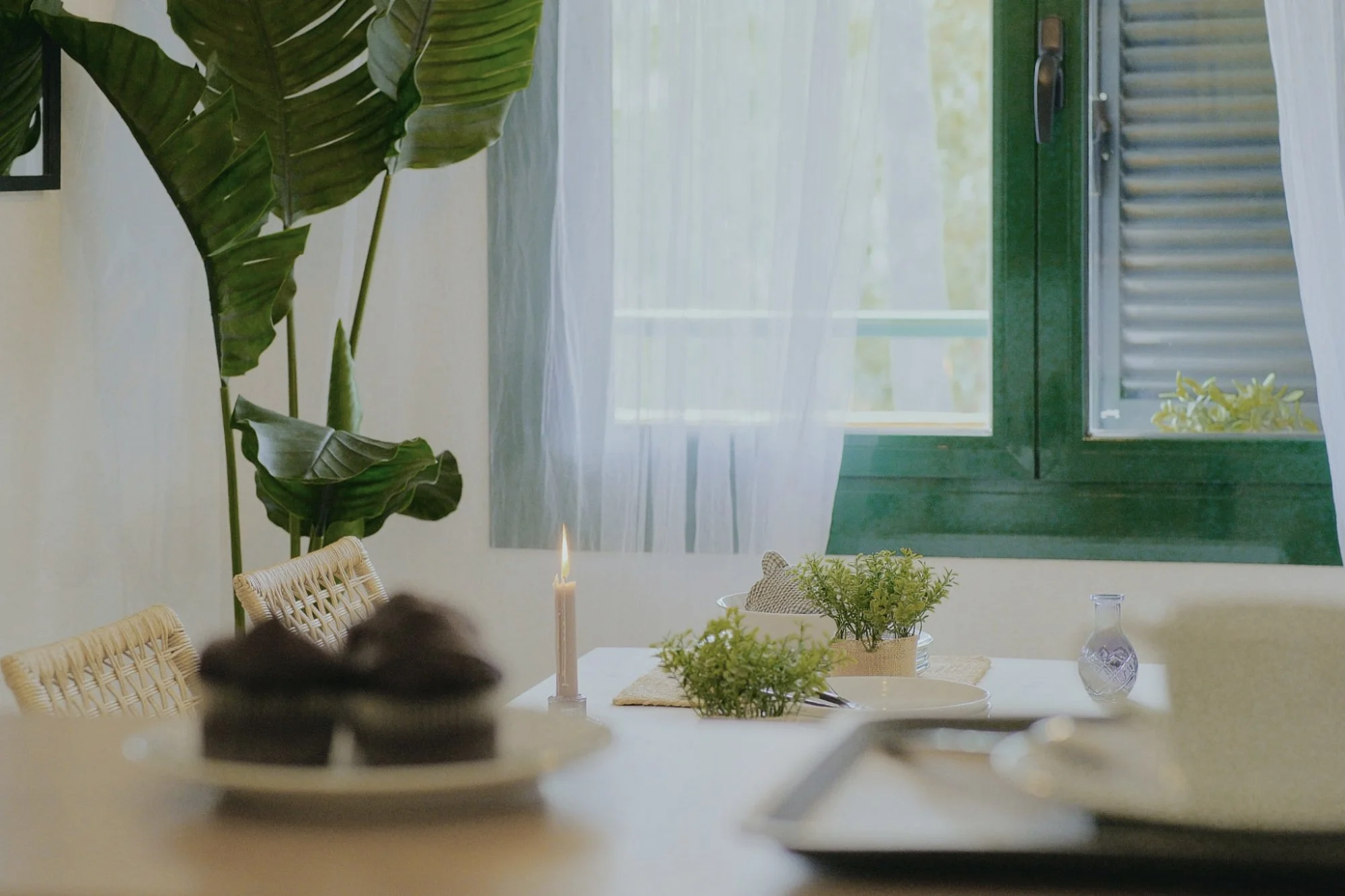 Mesa decorada con plantas, una vela encendida y jarrones transparentes. Fondo con ventana y cortinas blancas.