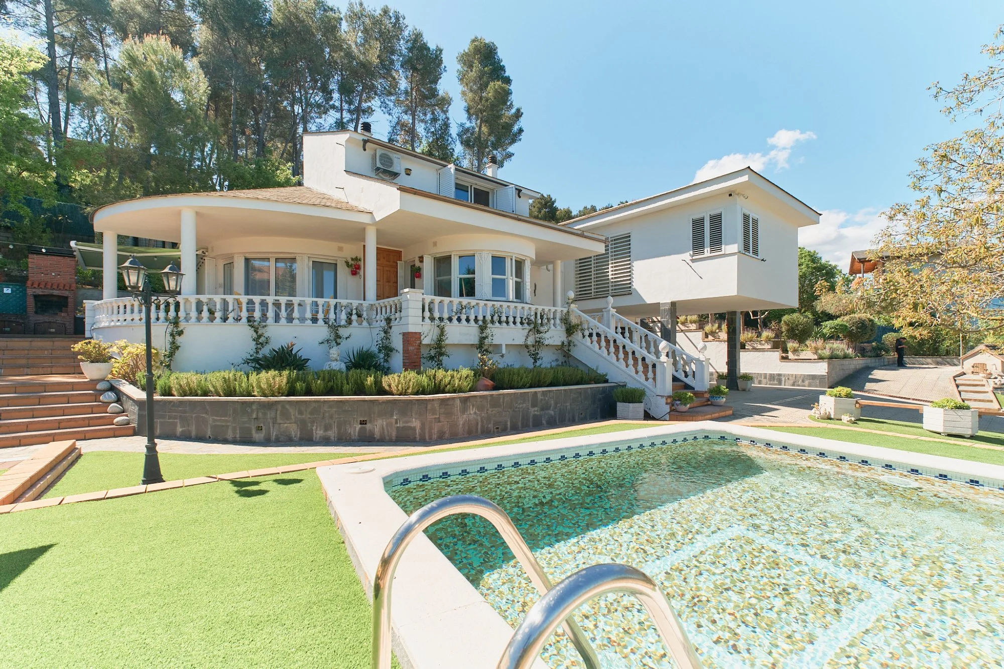 Casa moderna blanca con jardín y piscina en un entorno de árboles y cielo despejado.