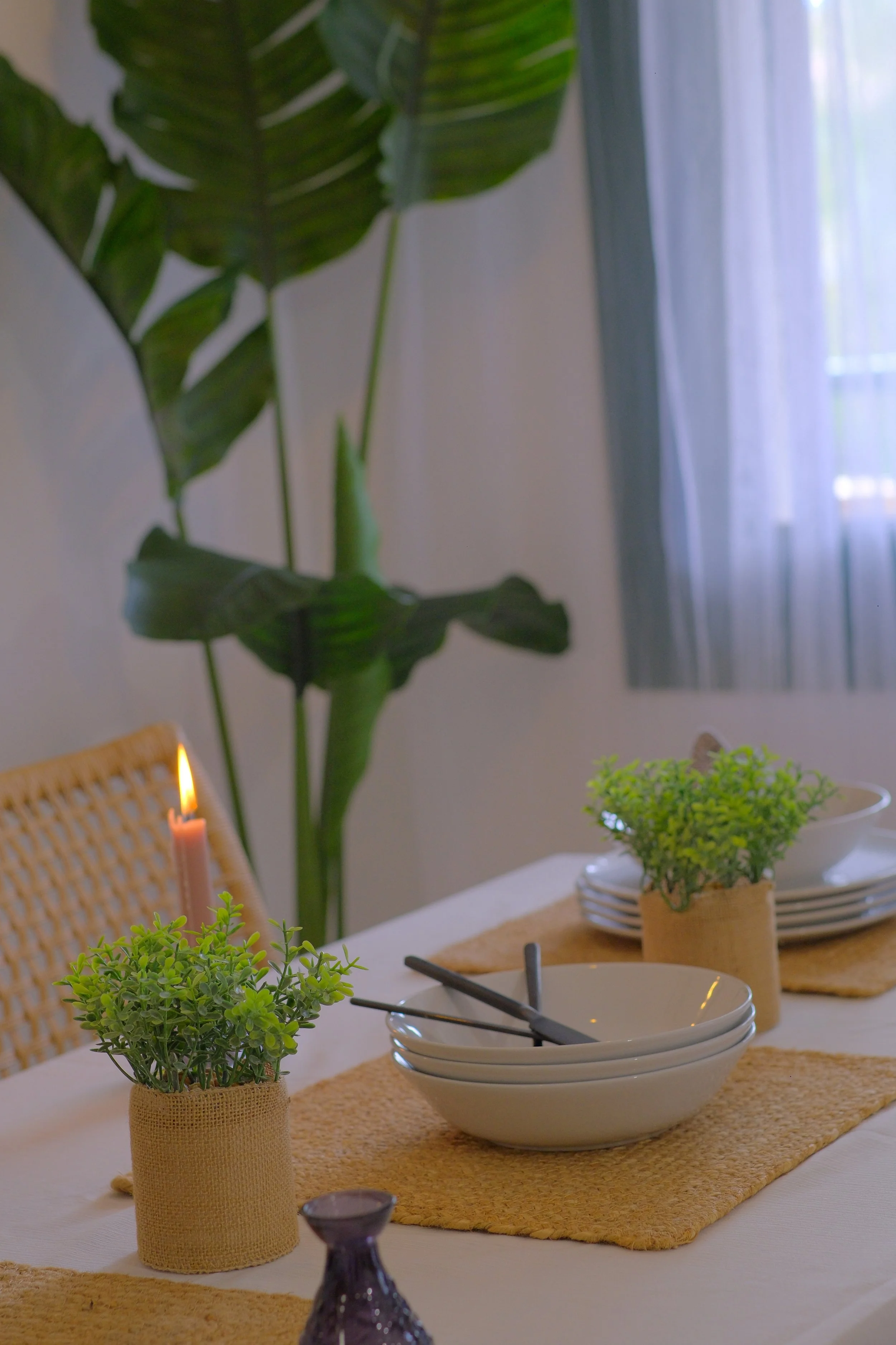 Mesa decorada con varias plantas en macetas de tela, platos blancos apilados, tazones, cubiertos y una vela en un candelabro, en un ambiente con cortinas blancas y una planta grande en el fondo.