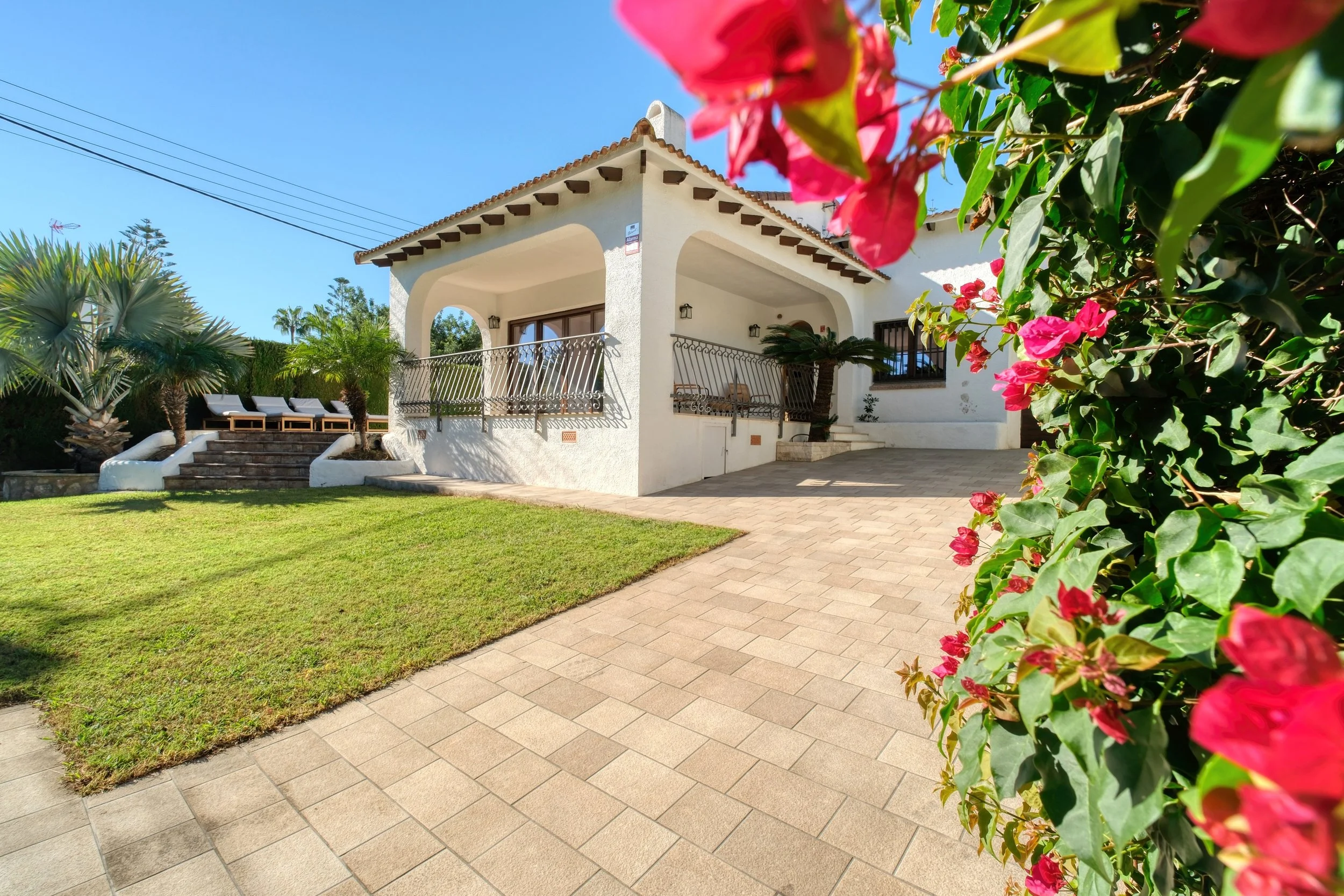 Casa blanca de estilo mediterráneo con balcón y jardineras, rodeada de plantas tropicales y flores rosadas, en un día soleado con cielo azul.