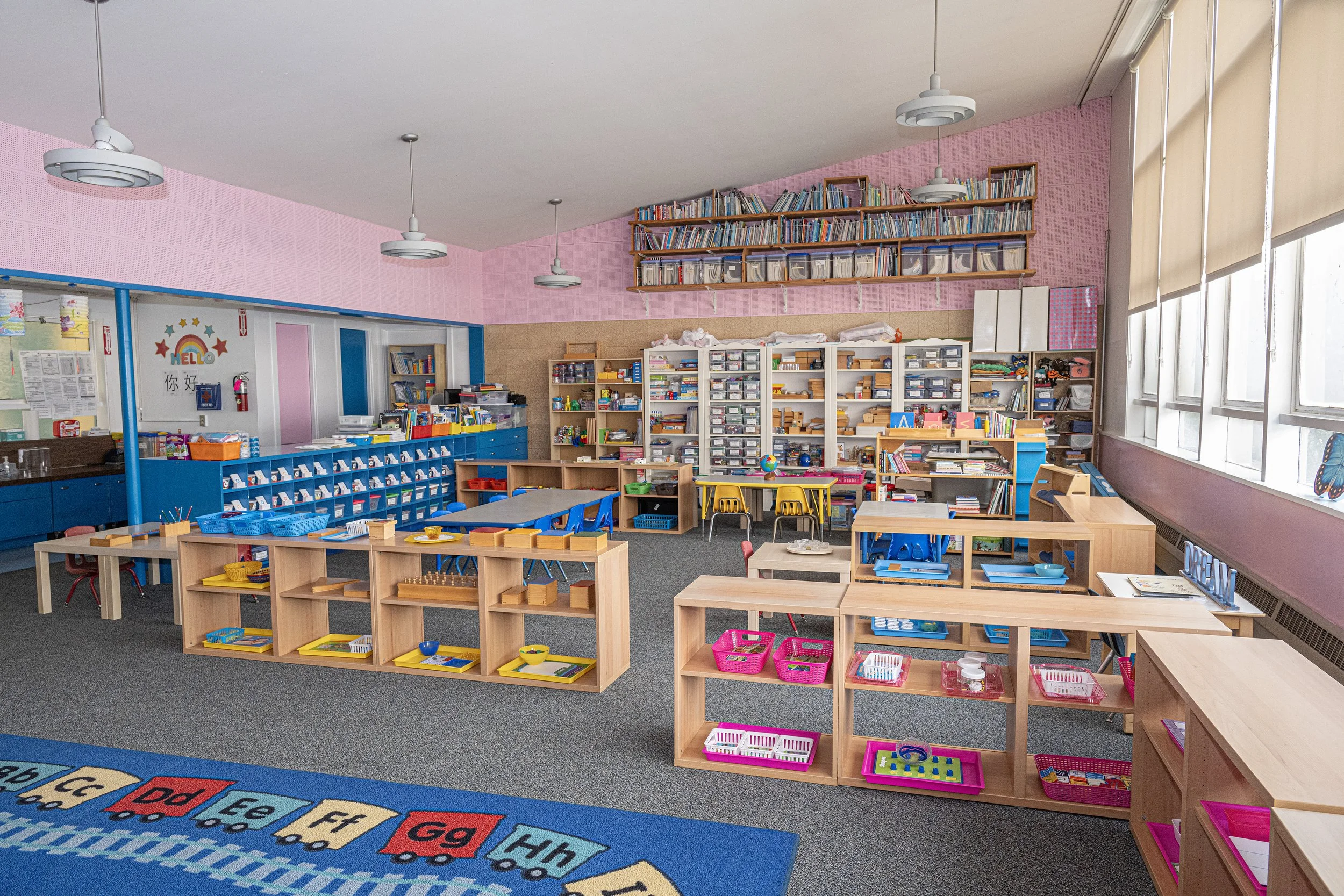 Colorful classroom with organized shelves of books and educational materials, small tables and chairs, and a large window letting in natural light.