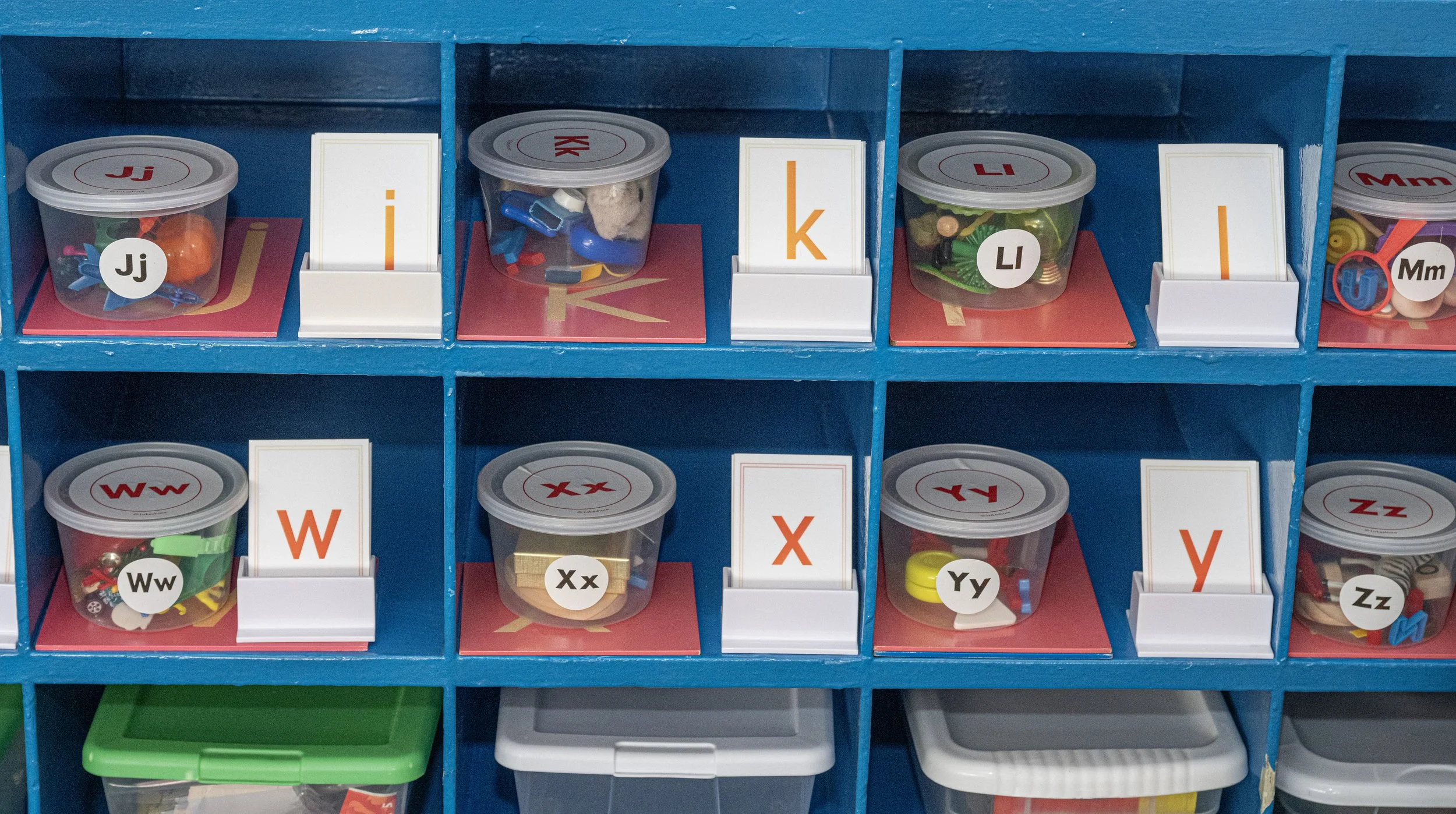 Blue cubby shelves with small clear containers labeled with letters and containing various small items and toys in a classroom setting.