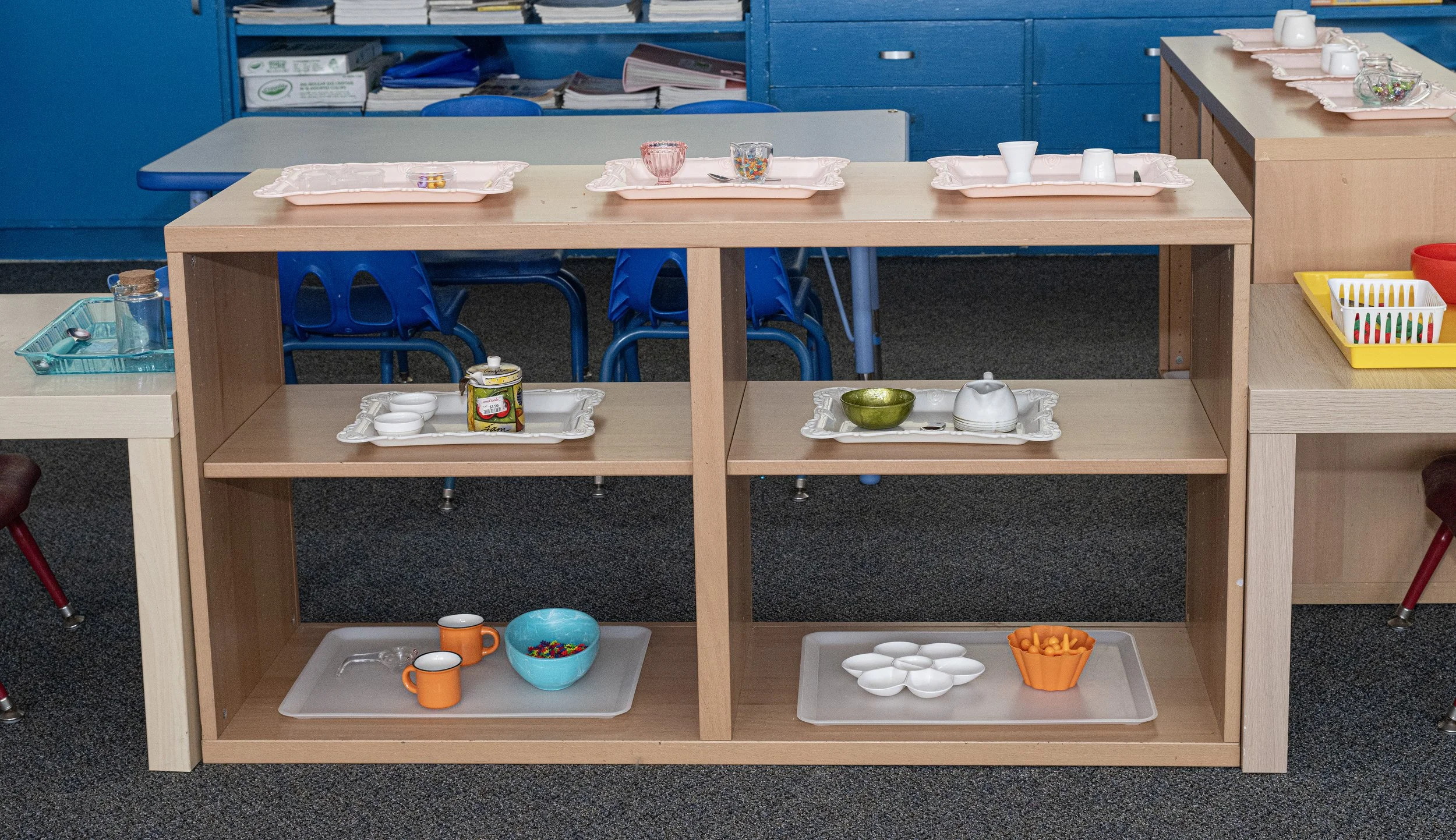 A classroom setting with several small tables, each with colorful trays, cups, bowls, and small objects for children's activities, and a wooden shelf in front holding additional items.