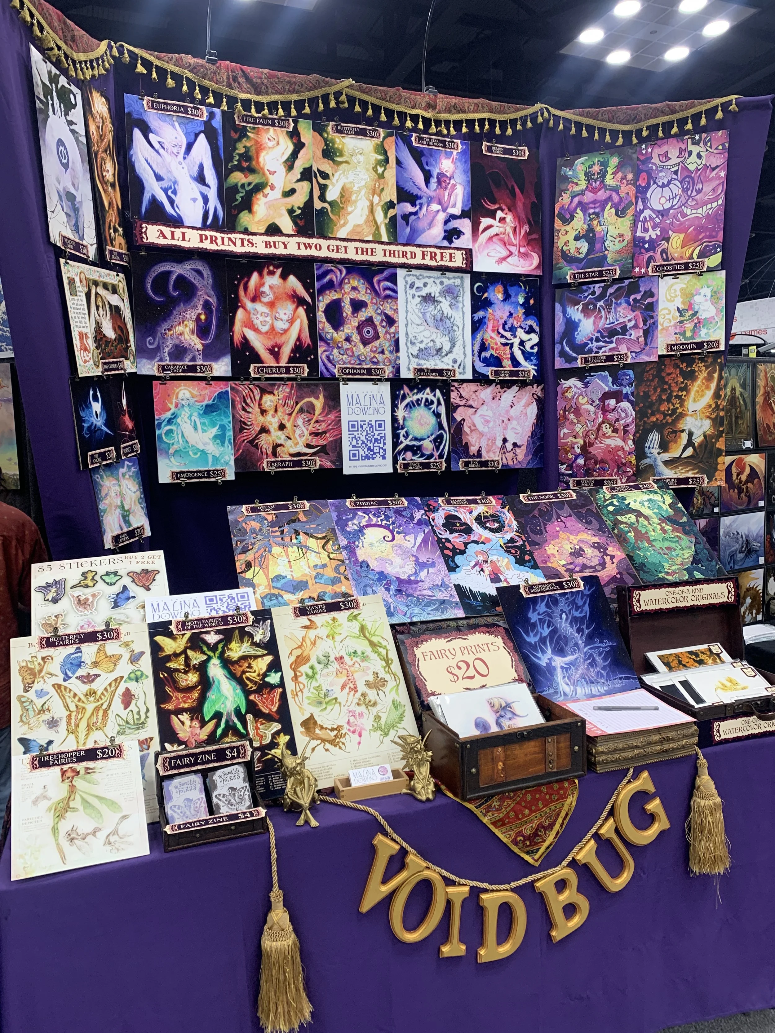 Purple and gold booth displaying various fantasy-themed art prints, stickers, and watercolor originals. “VOIDBUG” banner.