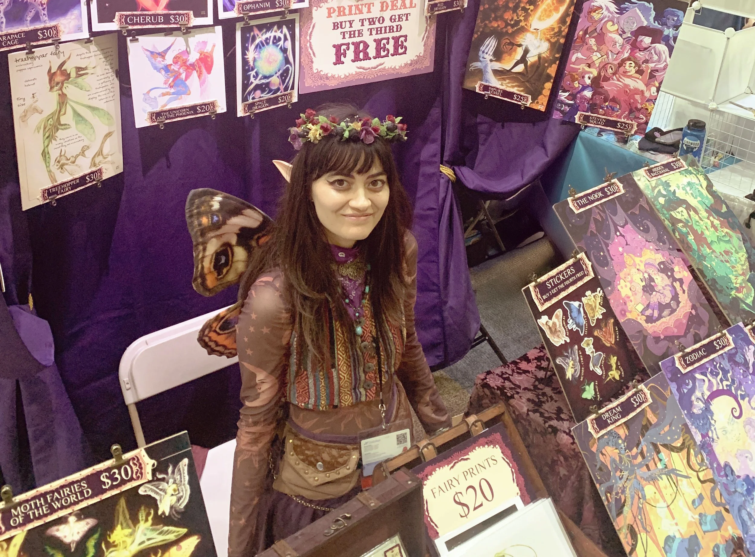 A woman dressed as a fairy with butterfly wings, a floral crown, and elf ears standing at a booth displaying fantasy-themed prints and stickers at a convention.
