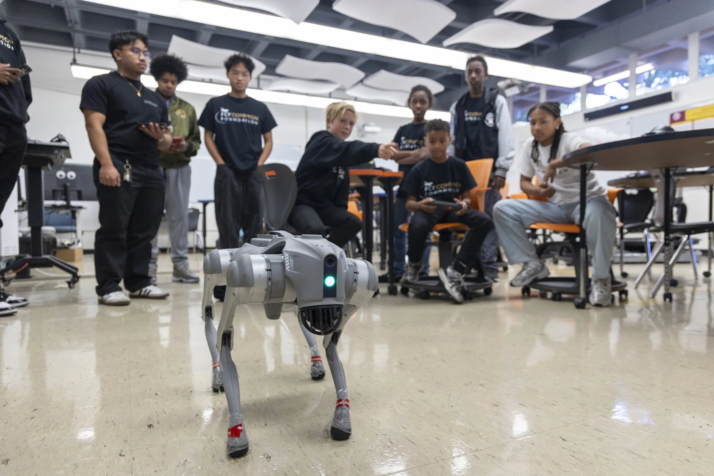 A group of young people in a classroom or laboratory setting, gathered around a person demonstrating a robot dog. The robot dog is gray with a glowing green light on its front, and the group appears attentive and engaged.