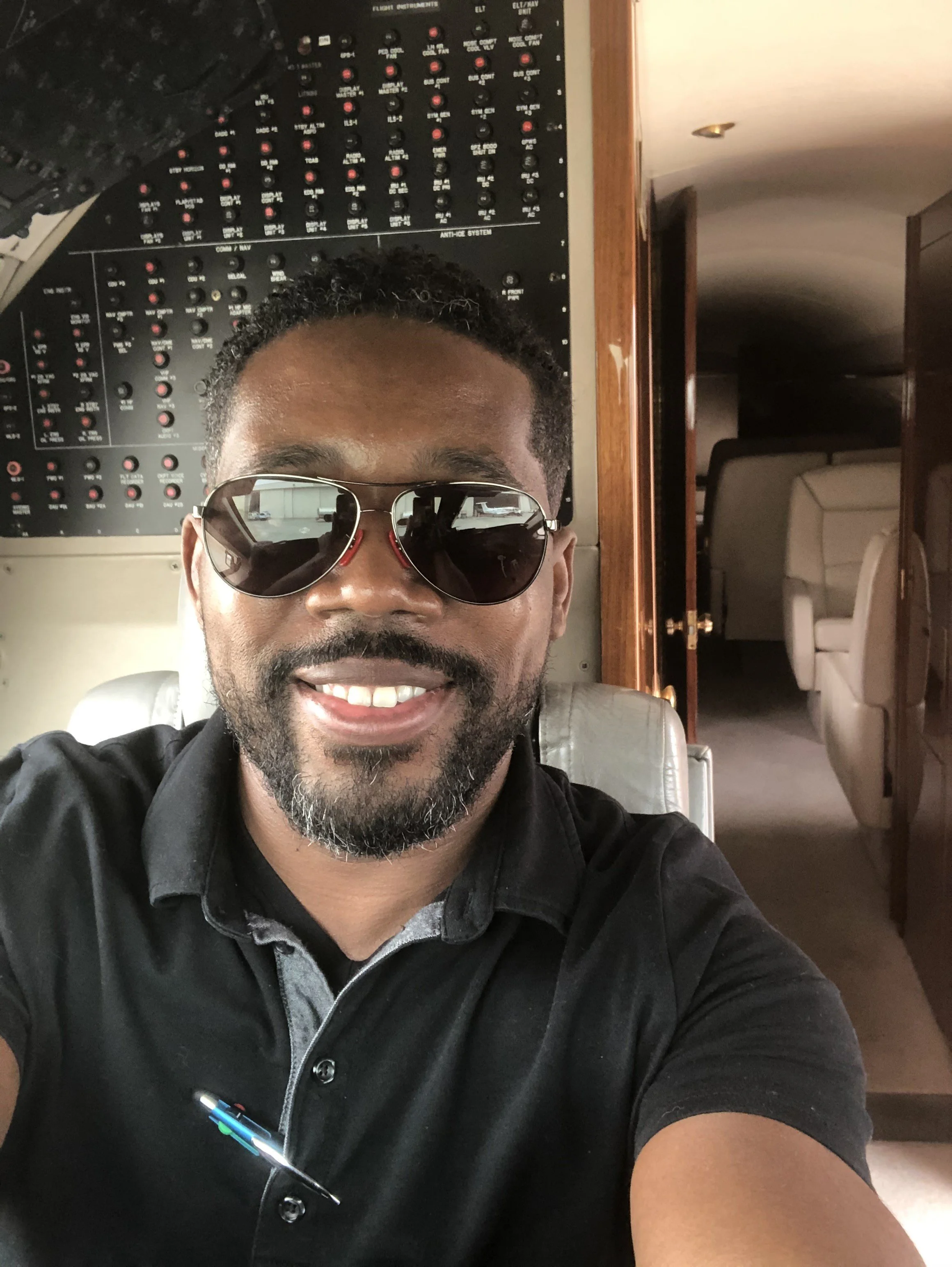 A smiling man with glasses taking a selfie inside an aircraft cockpit, with controls and a door leading to a passenger cabin in the background.