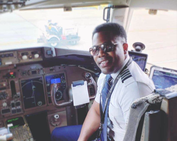 Pilot sitting in the cockpit of an airplane, wearing sunglasses and a uniform, smiling at the camera.