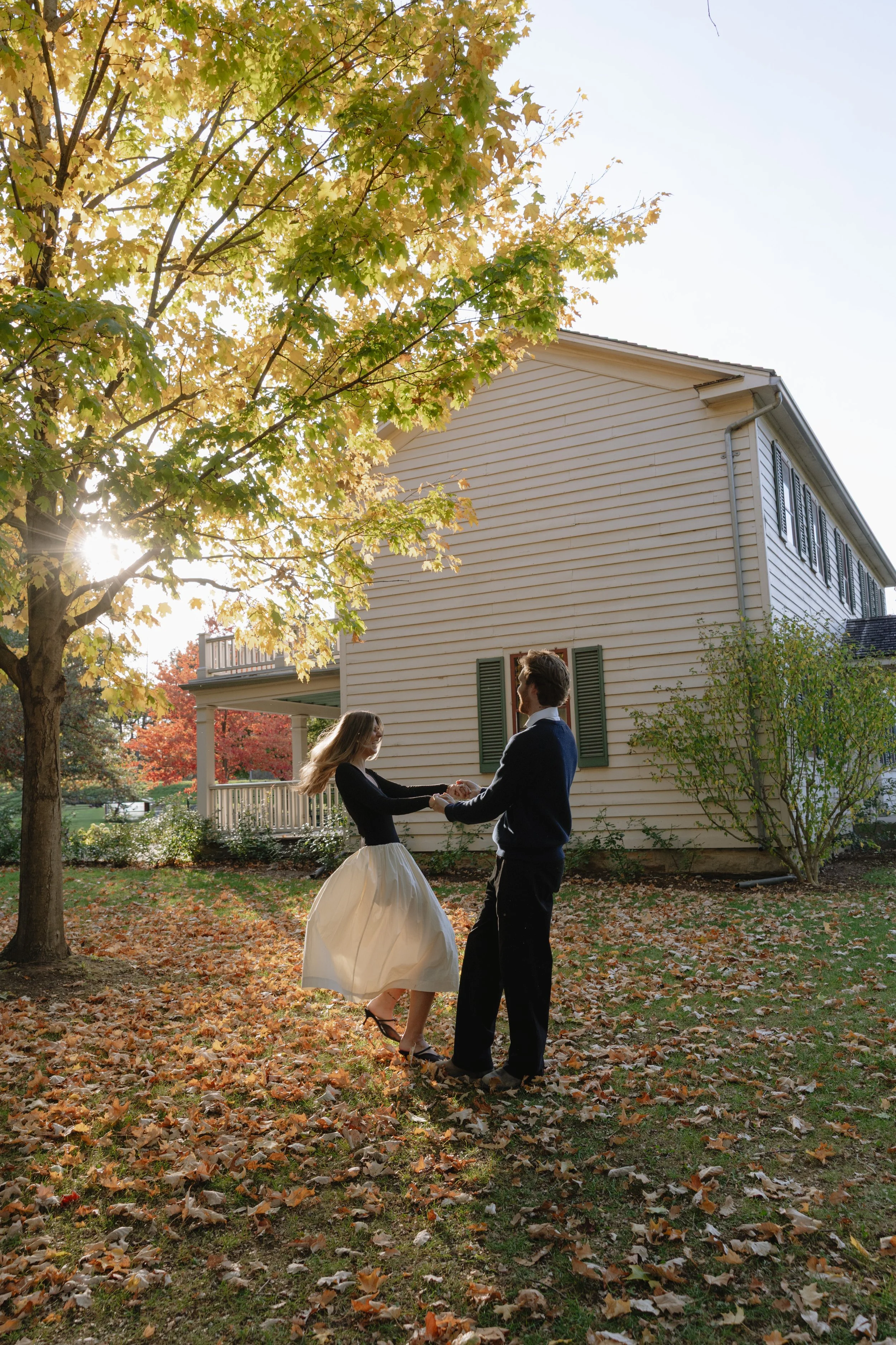 A couple dancing outdoors in the fall, surrounded by fallen leaves and trees with autumn foliage, in front of a white house with green shutters for their engagement session at Battlefield Park in Hamilton Ontario.
